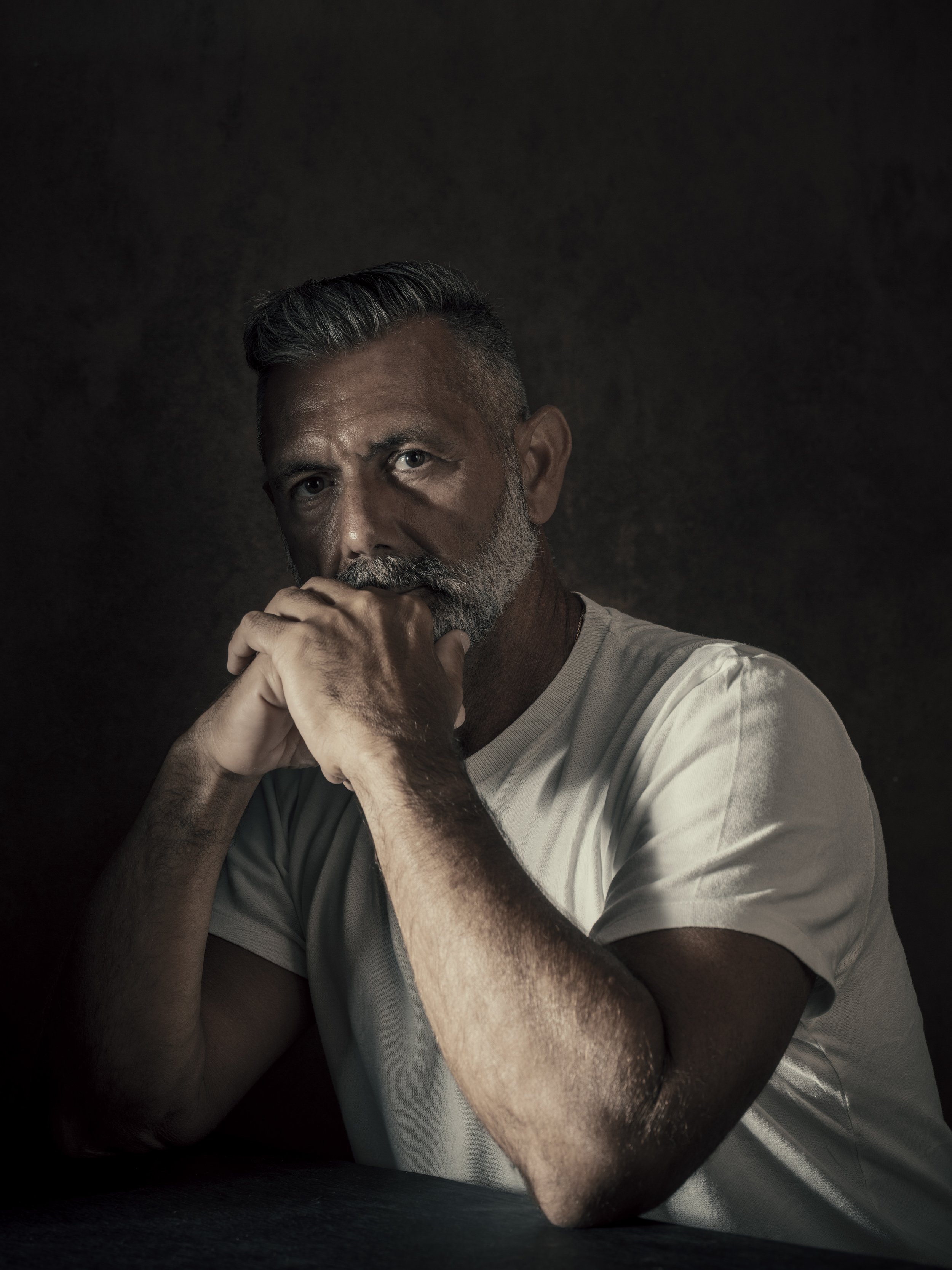 A close-up portrait of an older man with gray hair and beard, wearing a beige shirt, resting his chin on his hands against a dark background.