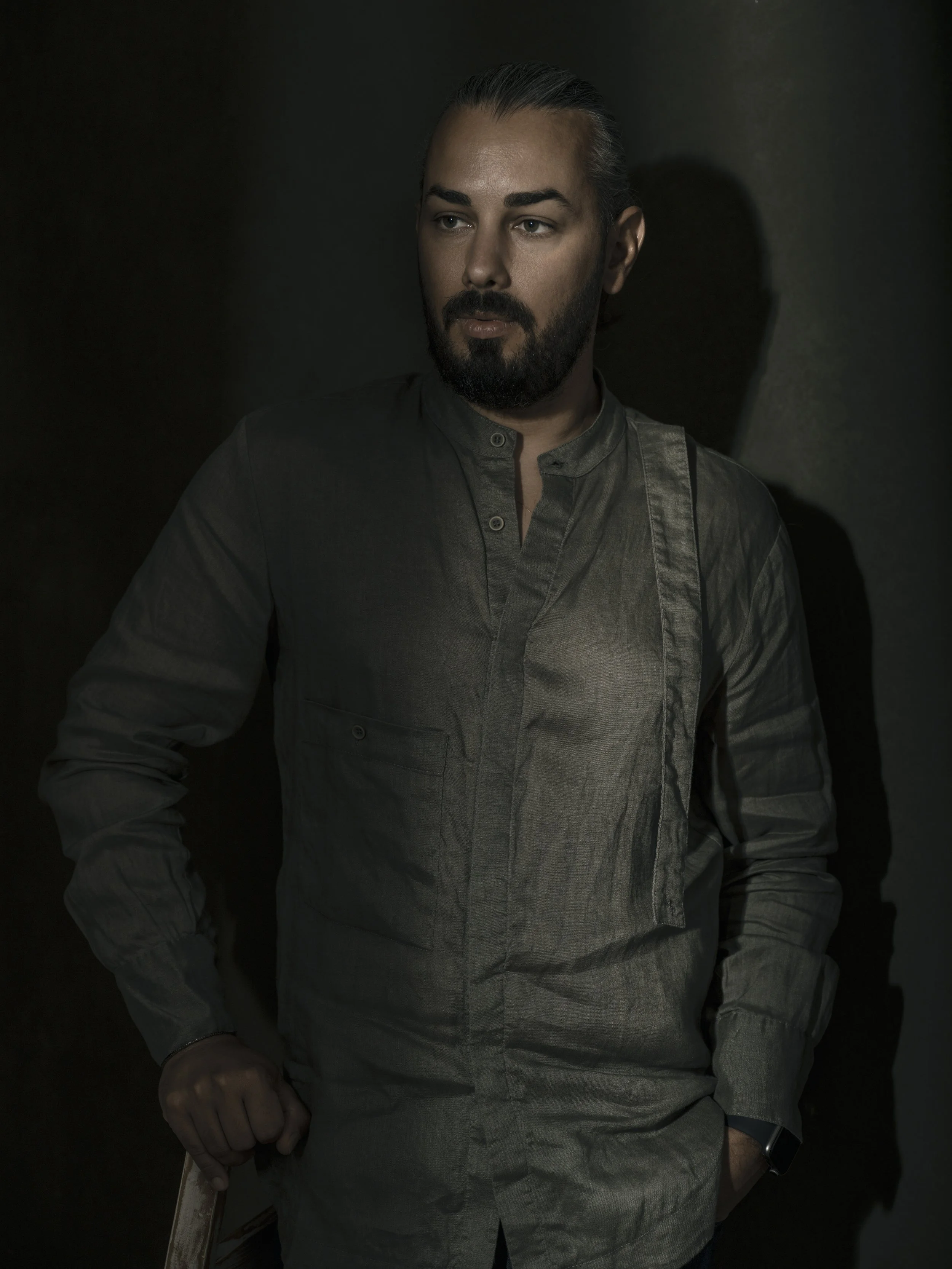 A man with dark hair, beard, and mustache, wearing a gray button-up shirt, seated against a dark background, looking to the side.