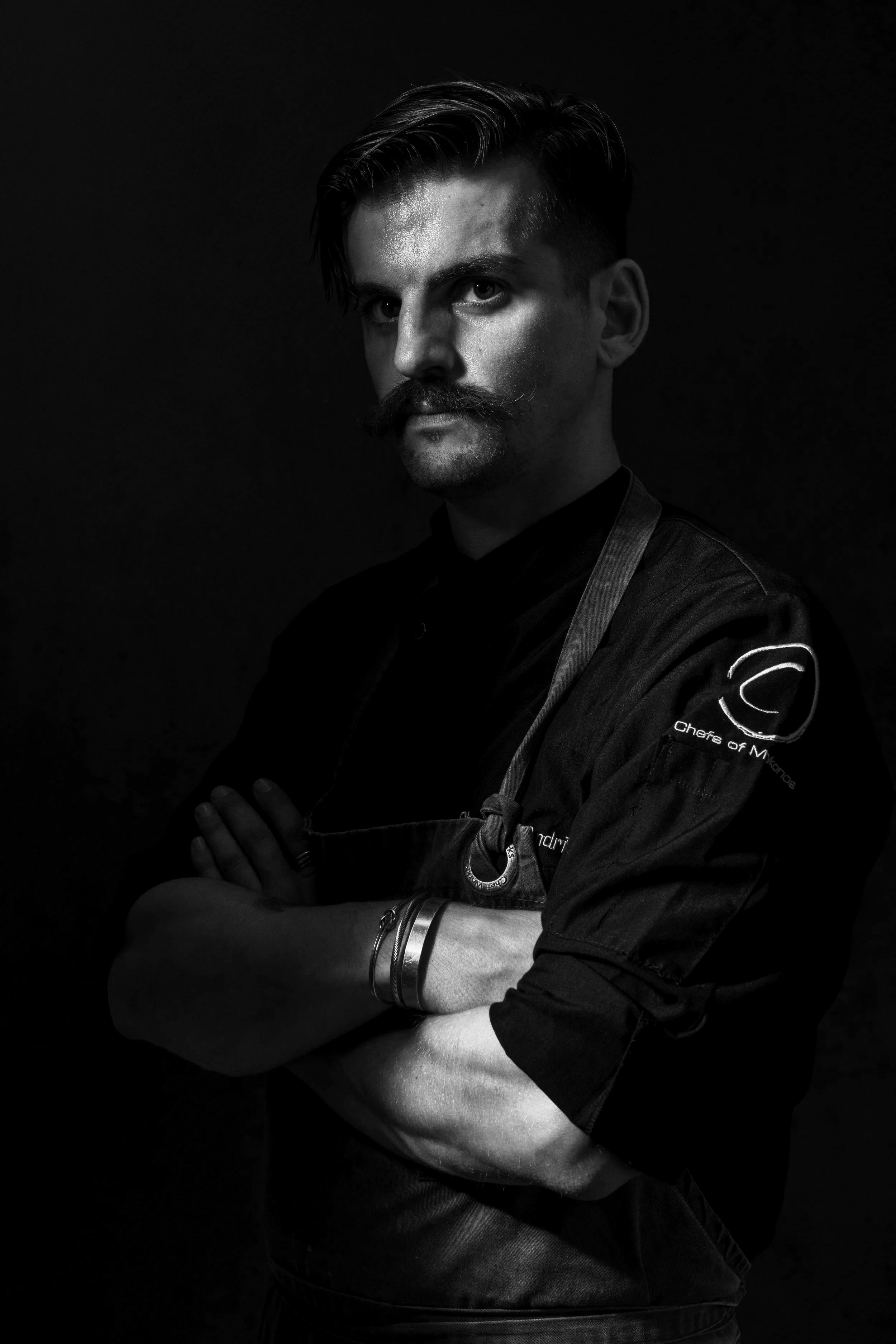 Black and white portrait of a man with a mustache and styled hair, wearing a chef's jacket with patches on the sleeve, standing with arms crossed against a dark background.