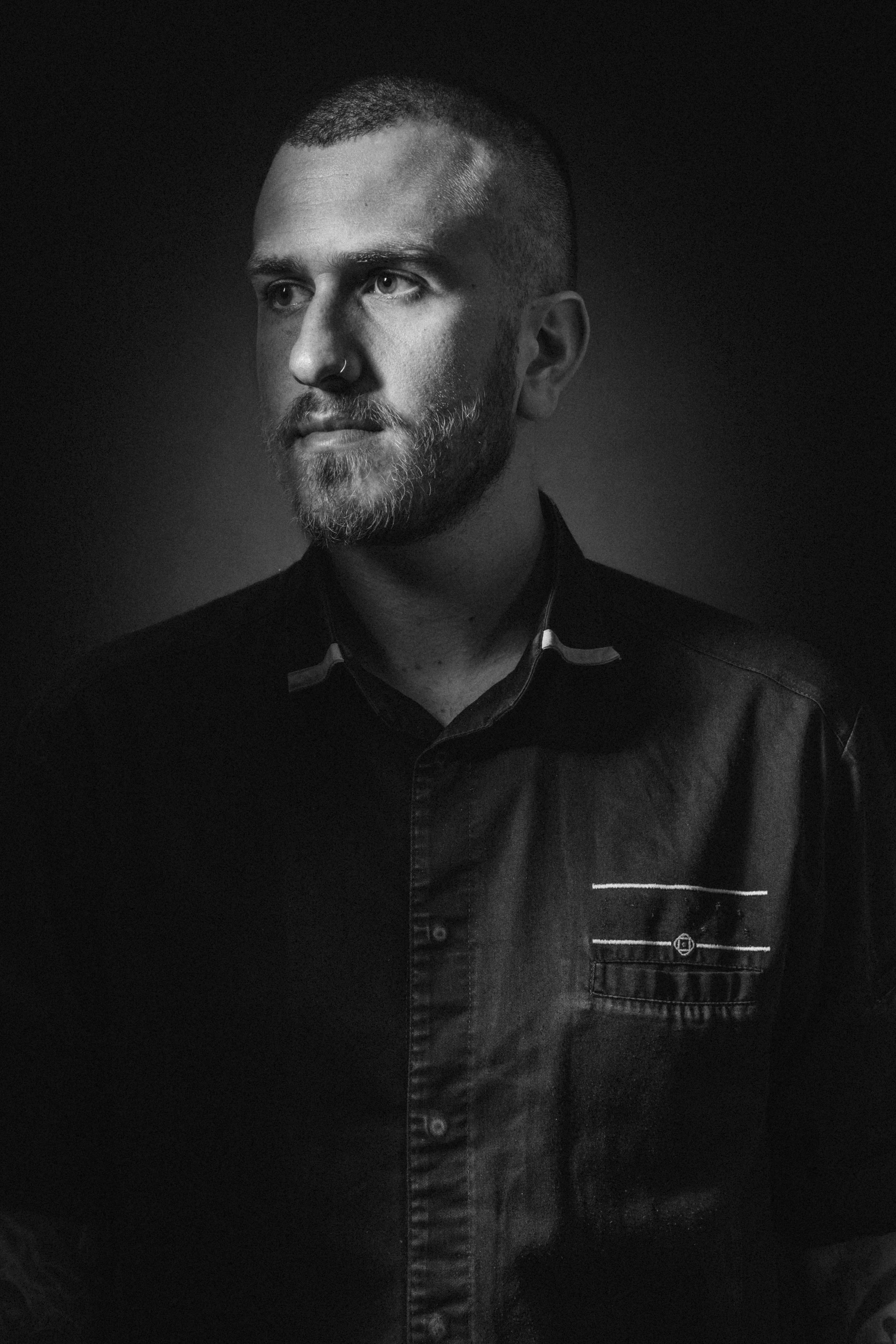Black and white portrait of a man with a beard, mustache, short hair, and a nose ring, wearing a collared shirt with a pocket and pattern, against a dark background.