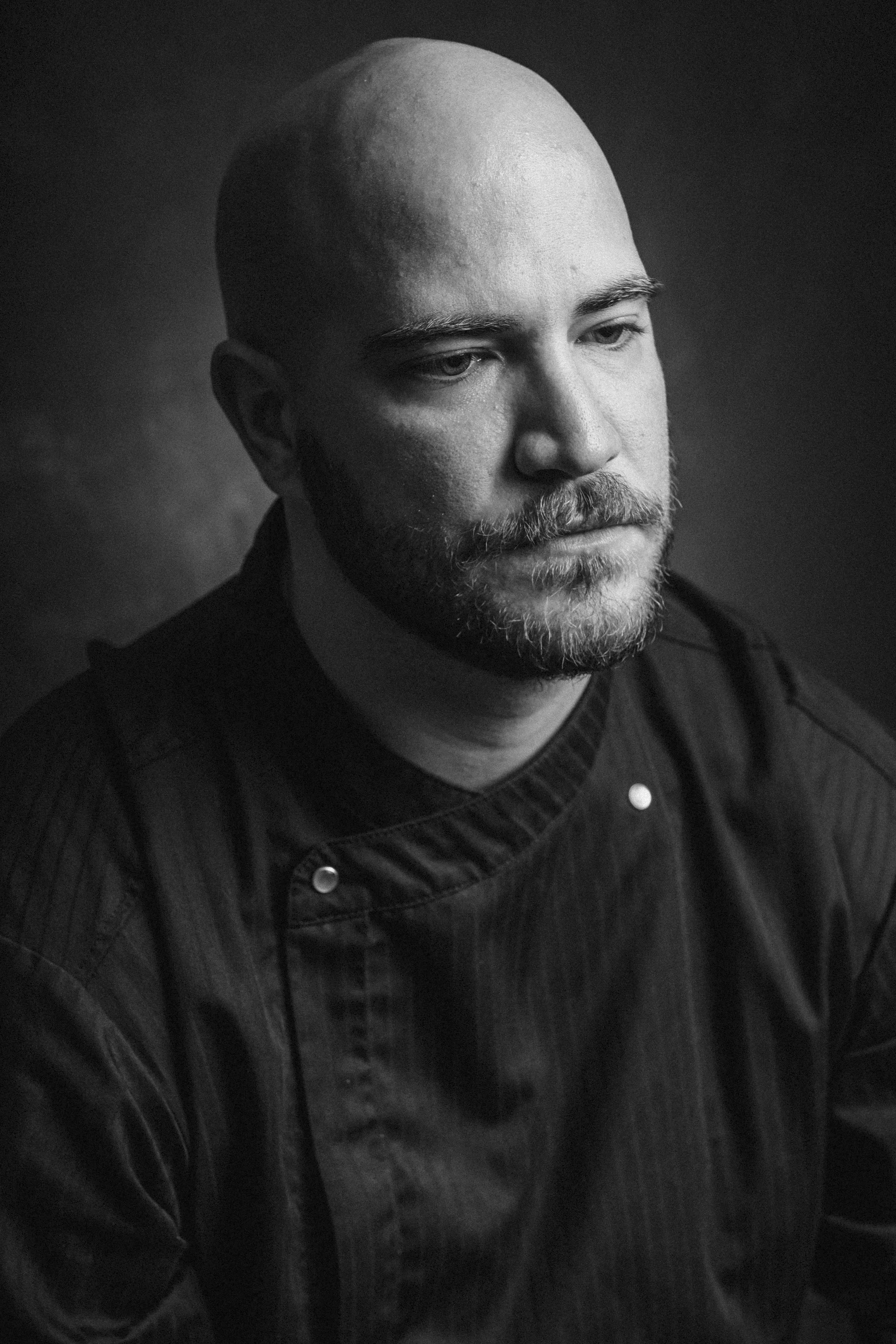 A black and white portrait of a man with a beard and shaved head, wearing a dark shirt or jacket.