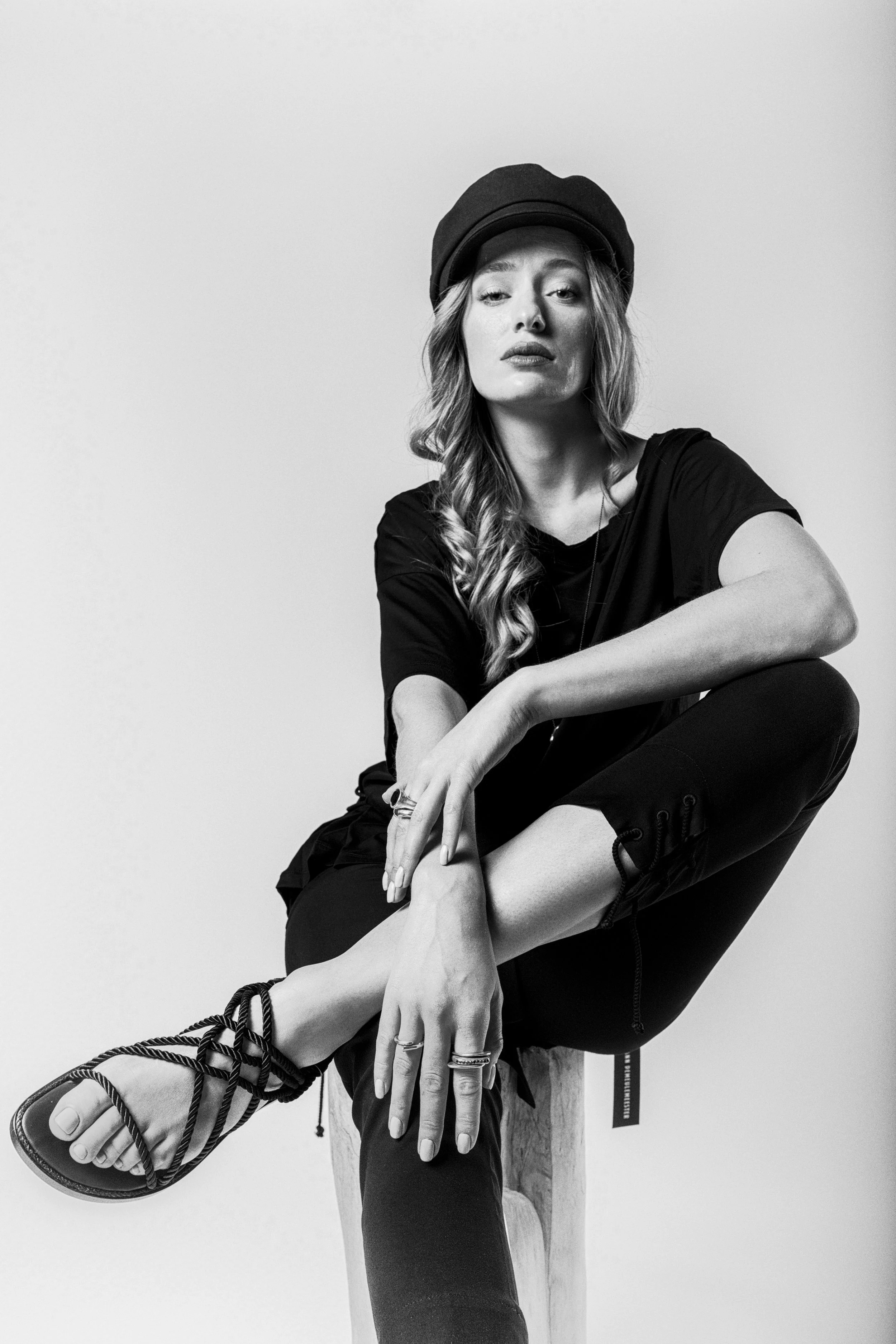 Black and white portrait of a woman with long wavy hair, wearing a beret, a casual black t-shirt, and pants. She is sitting with her legs crossed, one arm resting on her knee and the other hanging down, wearing rings and multiple bracelets. She gazes