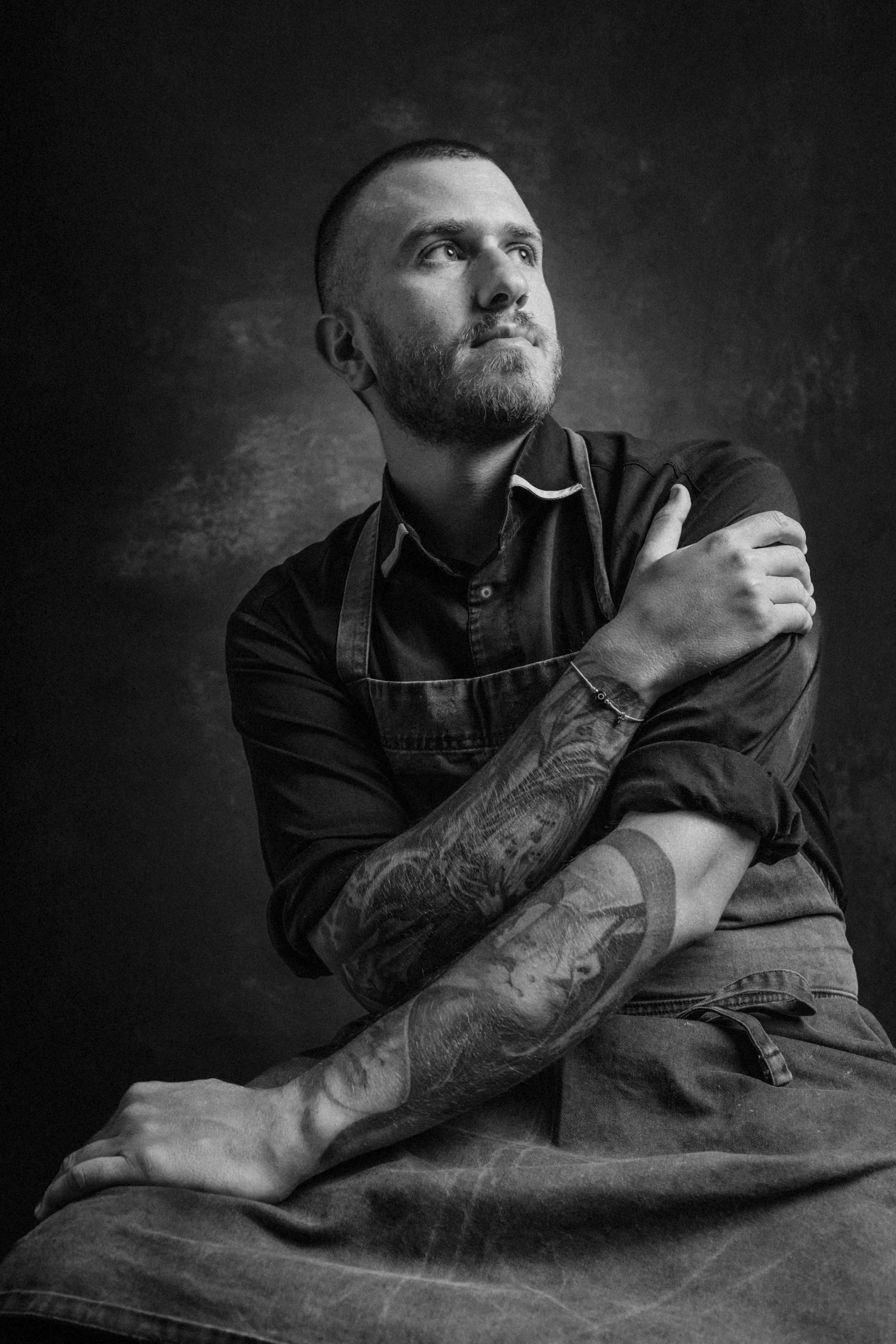 Black and white portrait of a bearded man with short hair looking upwards. He is wearing a dark shirt with a collar and an apron, possibly indicating he is a chef or artist. His arms are crossed, revealing detailed tattoos on one forearm. The backgro