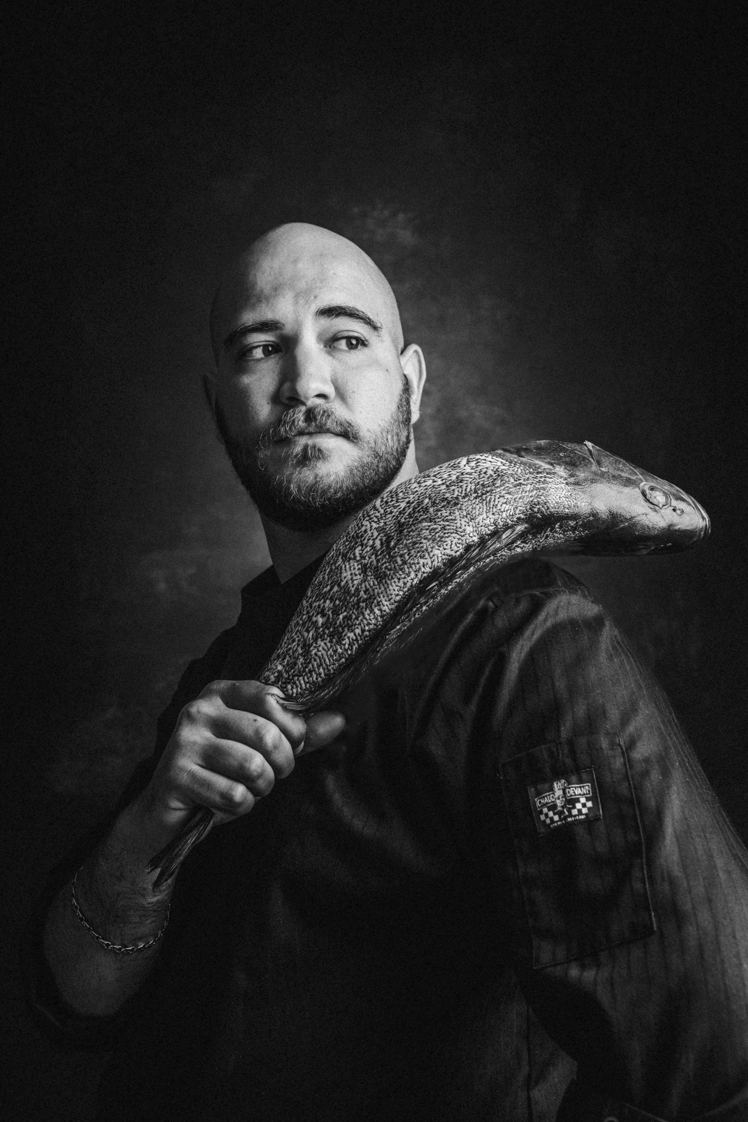 Black and white portrait of a man with a beard, holding a large fish over his shoulder, wearing a jacket with a logo patch. The background is dark and plain.