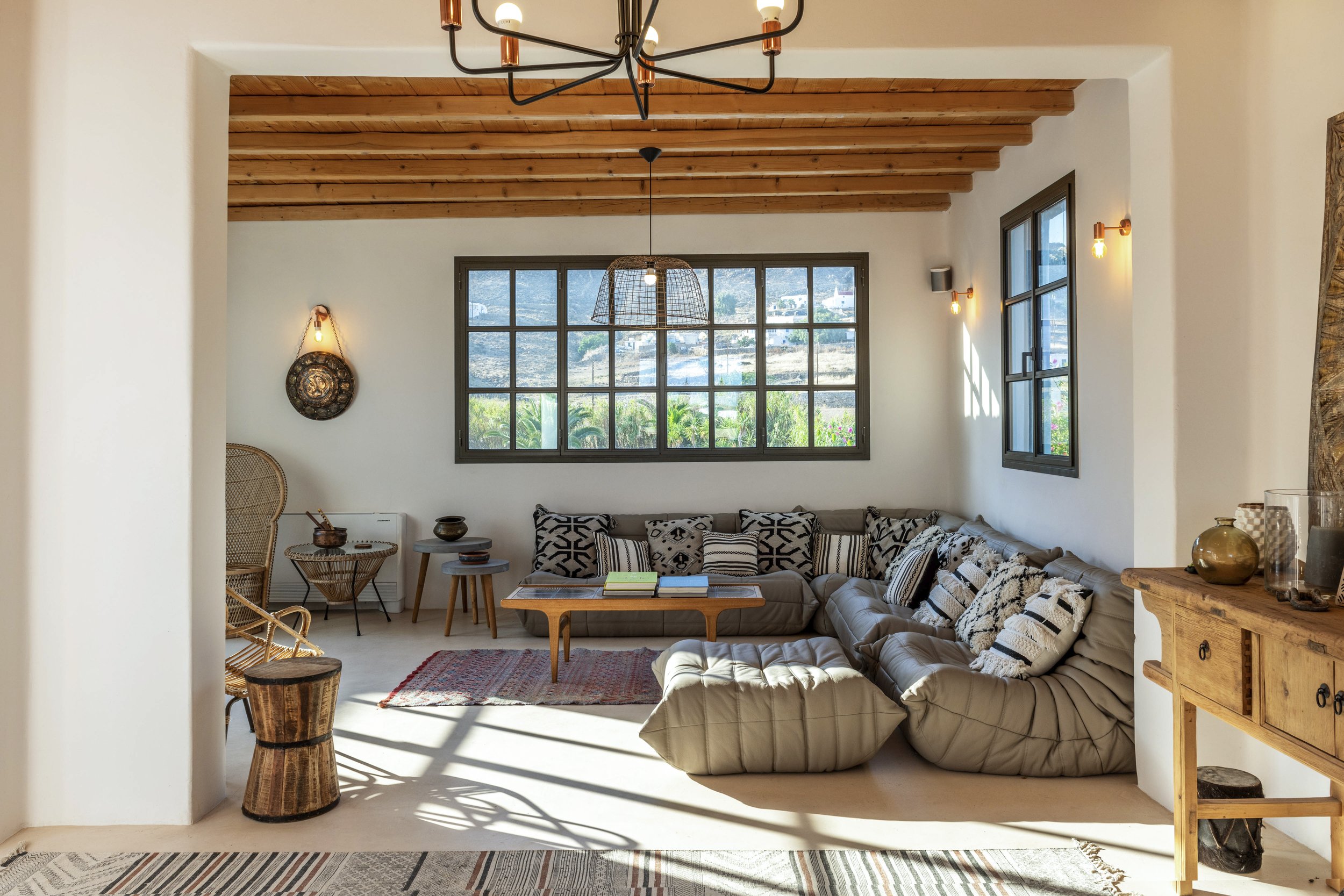 Living room with beige sectional sofa, black and white patterned pillows, wooden coffee table with books, windows showing an outdoor landscape, wooden ceiling beams, and various decorative elements.