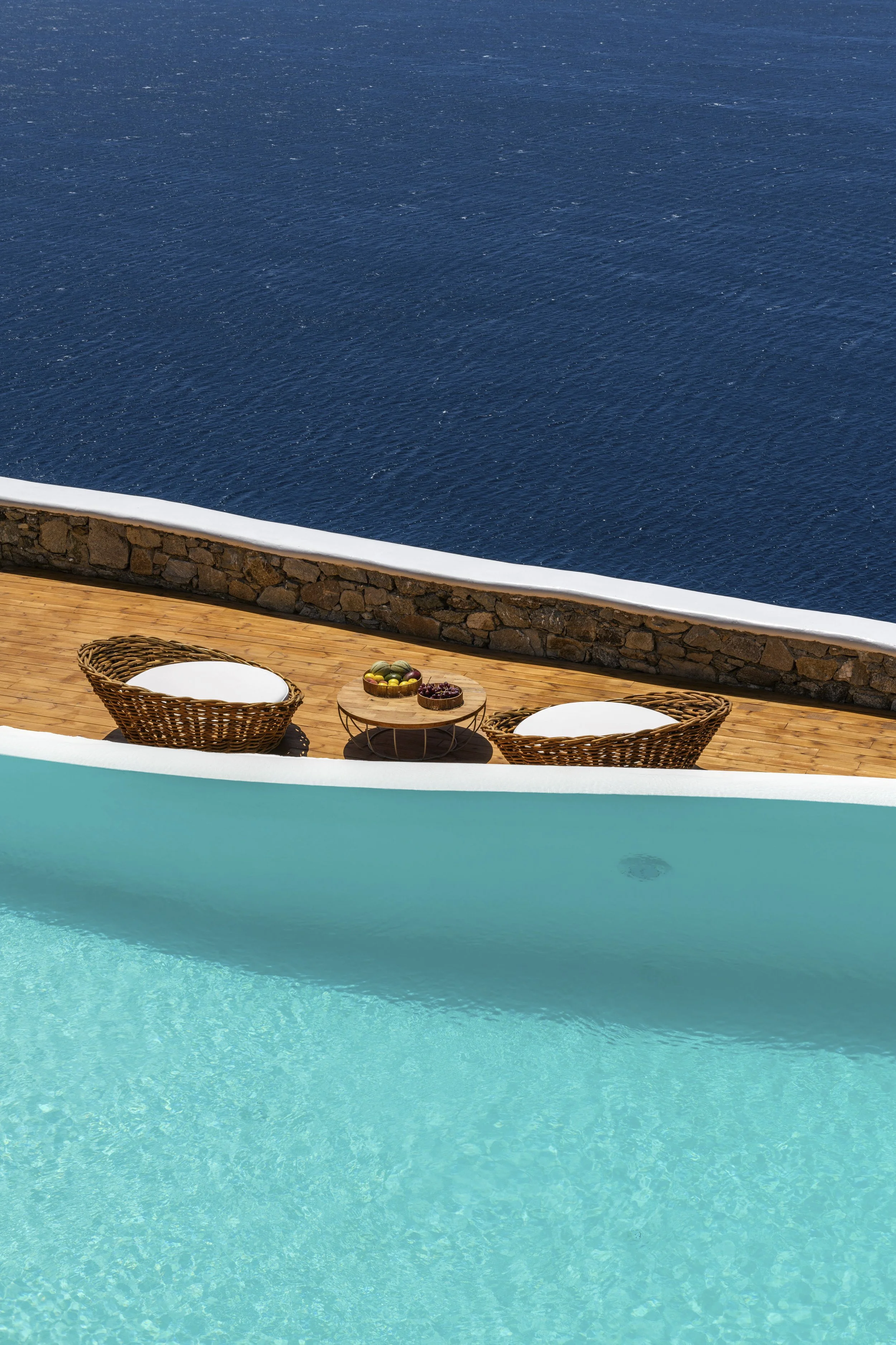 View of a swimming pool with a turquoise water surface, a wooden deck, two wicker chairs with white cushions, a small wooden table with a bowl of fruit, a stone wall, and the ocean in the background.