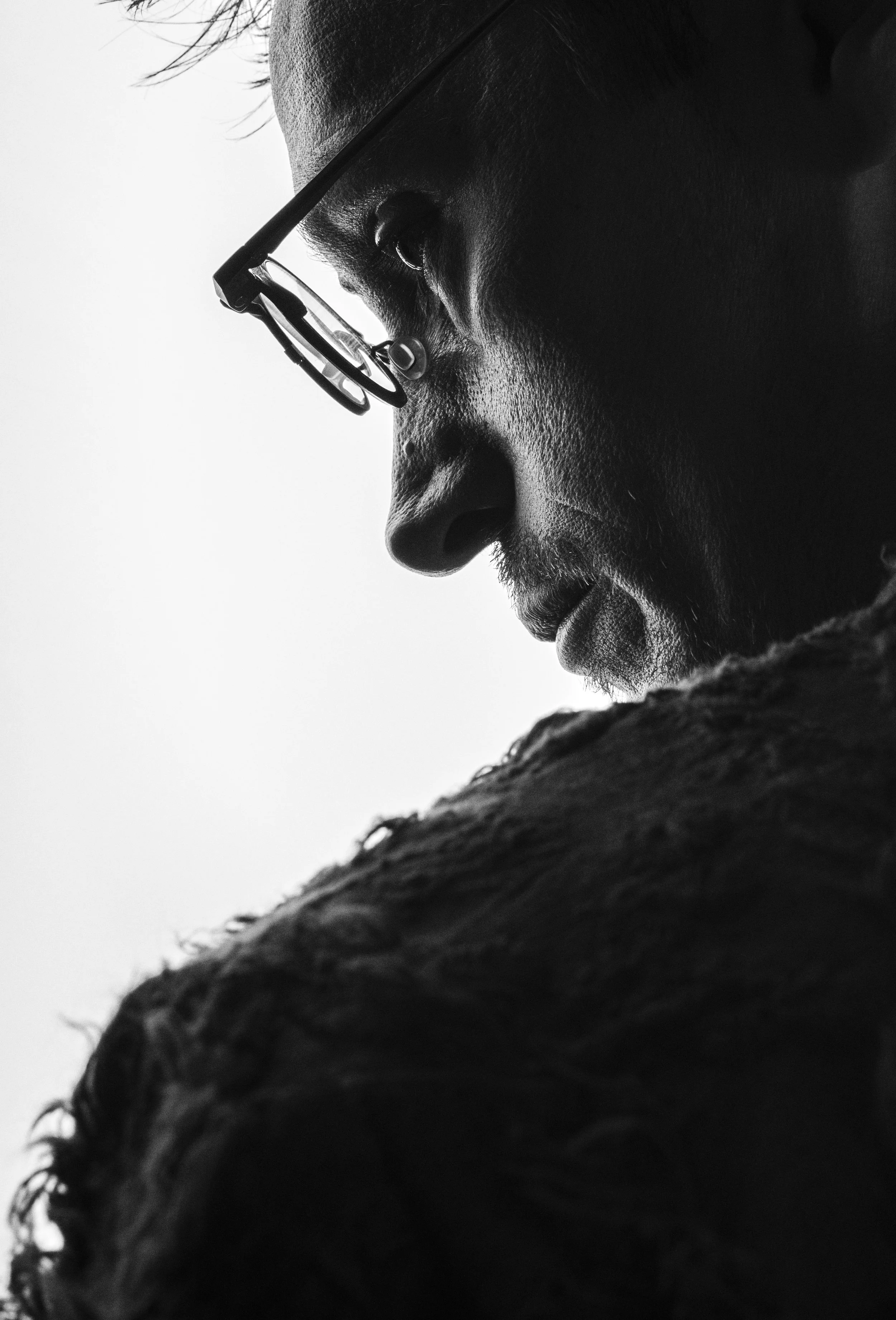 Close-up side profile of a person's face wearing glasses, with textured skin and short curly hair, in black and white.