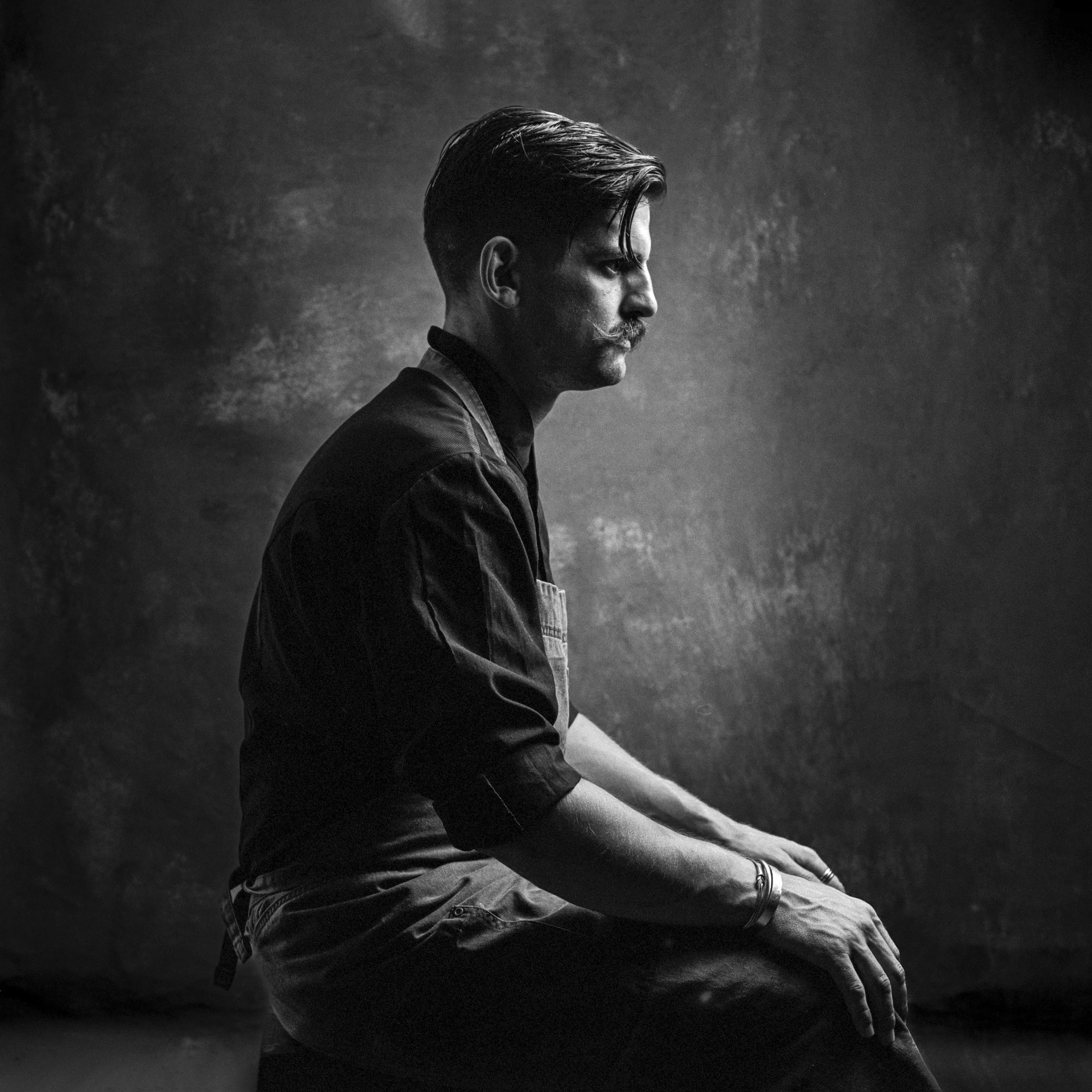 A black and white photo of a man with dark hair and a mustache, wearing a suit, looking to the side with a serious expression, against a dark, textured background.