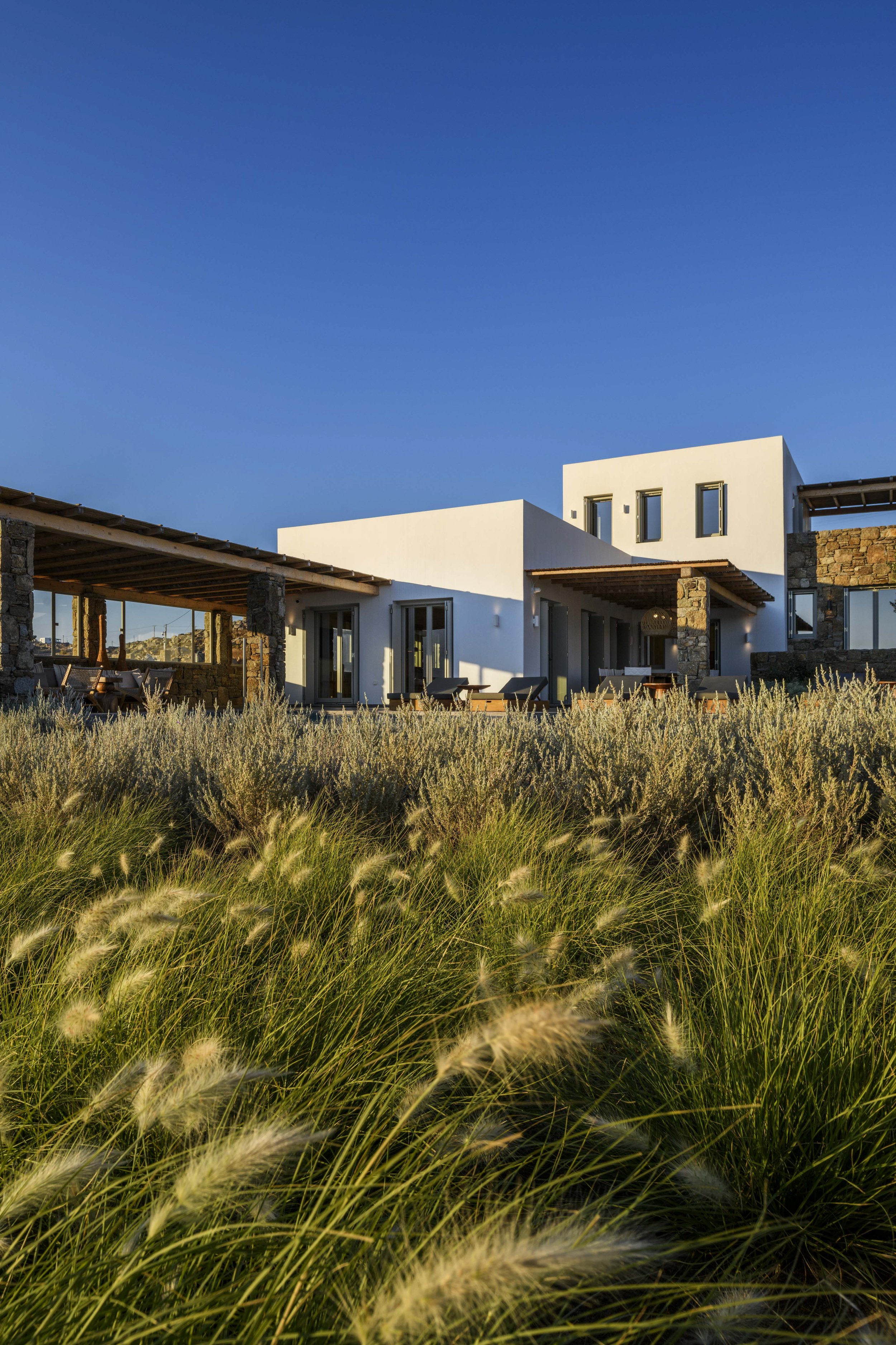 Modern white house with stone accents, multiple windows, and outdoor seating area, surrounded by tall grass under a clear blue sky.