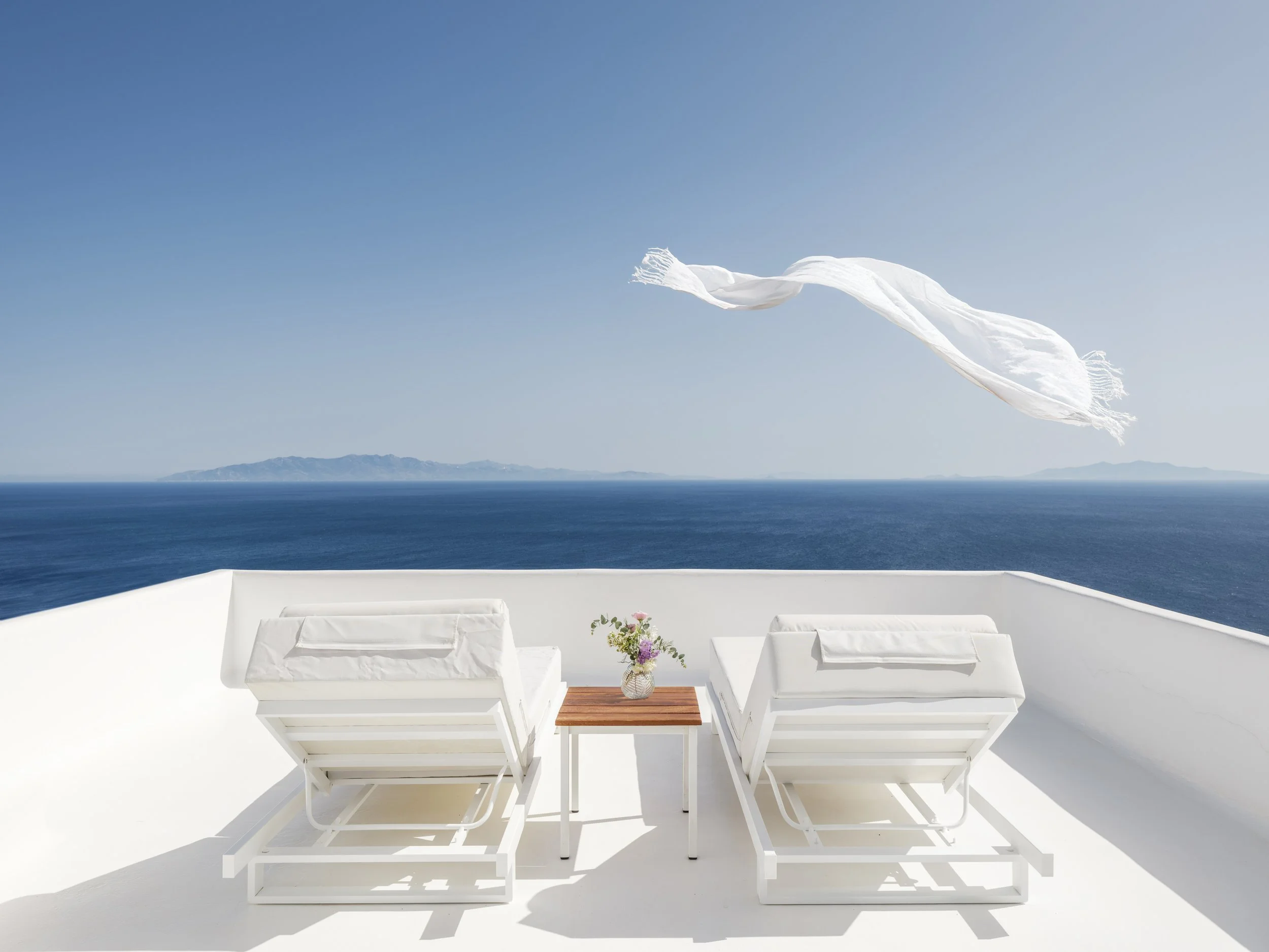 White outdoor terrace with two lounge chairs with white cushions, a small wooden table with a flower vase, overlooking the blue sea, a distant mountainous island, under a clear blue sky, with a white fabric windbreaker flowing.