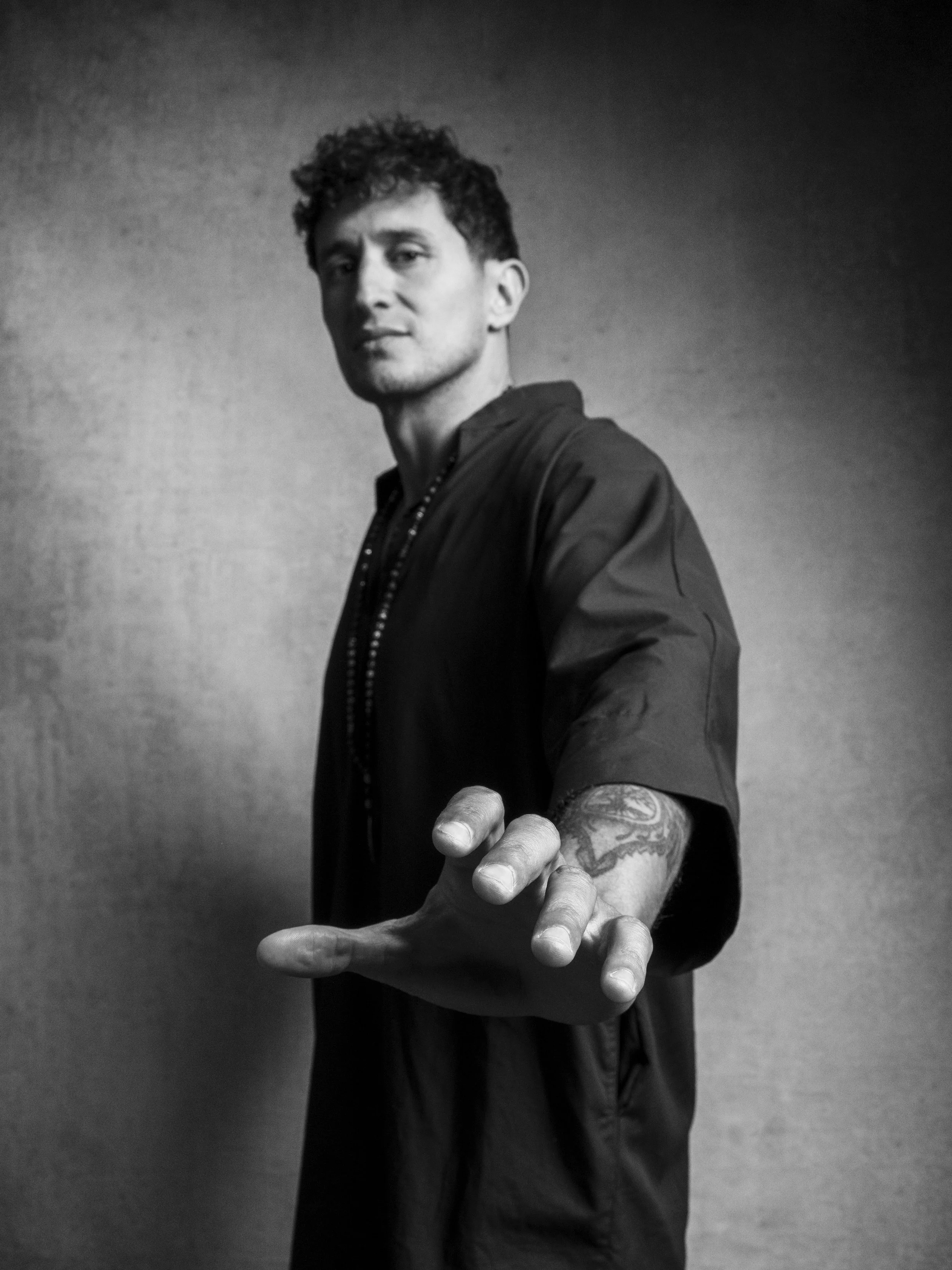 A black and white photo of a man with curly hair and tattoos on his right arm extending his hand toward the camera.