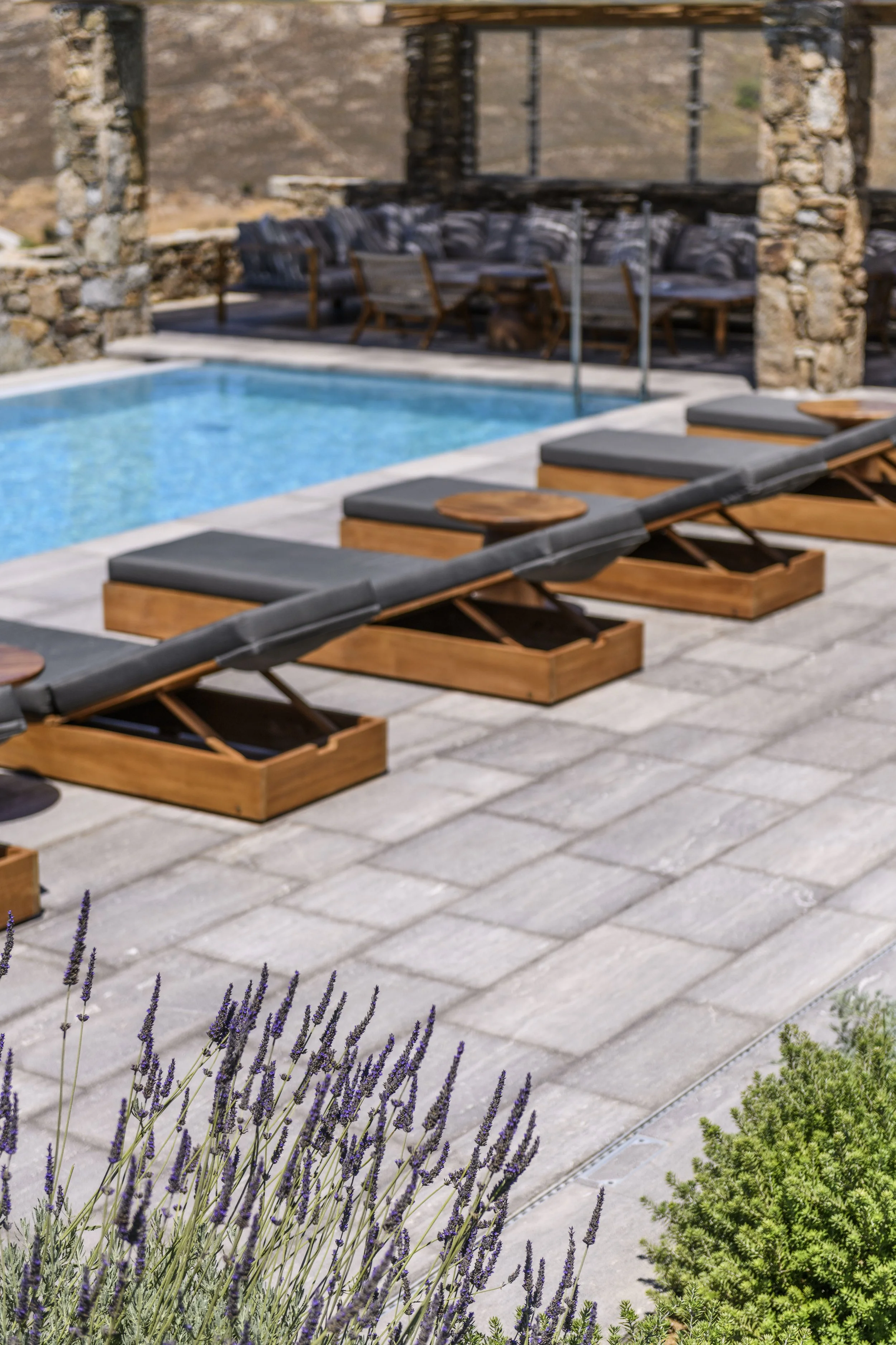 Outdoor patio with lounge chairs, a swimming pool, stone pillars, and mountain scenery in the background. Purple lavender plants in the foreground.