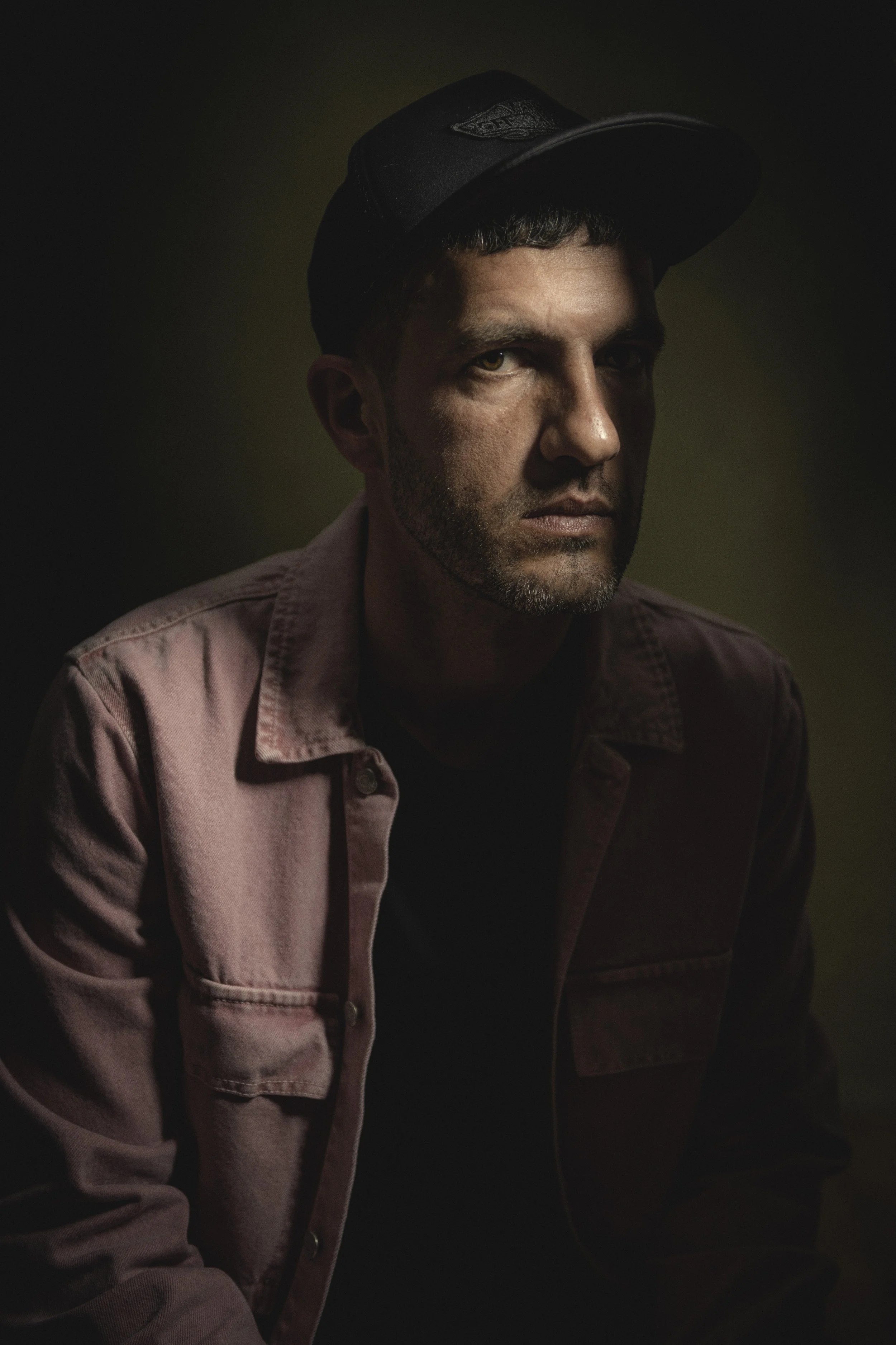 A man with short dark hair and facial hair wearing a black cap and a light pink denim jacket, sitting in a dark environment with a faint greenish background.