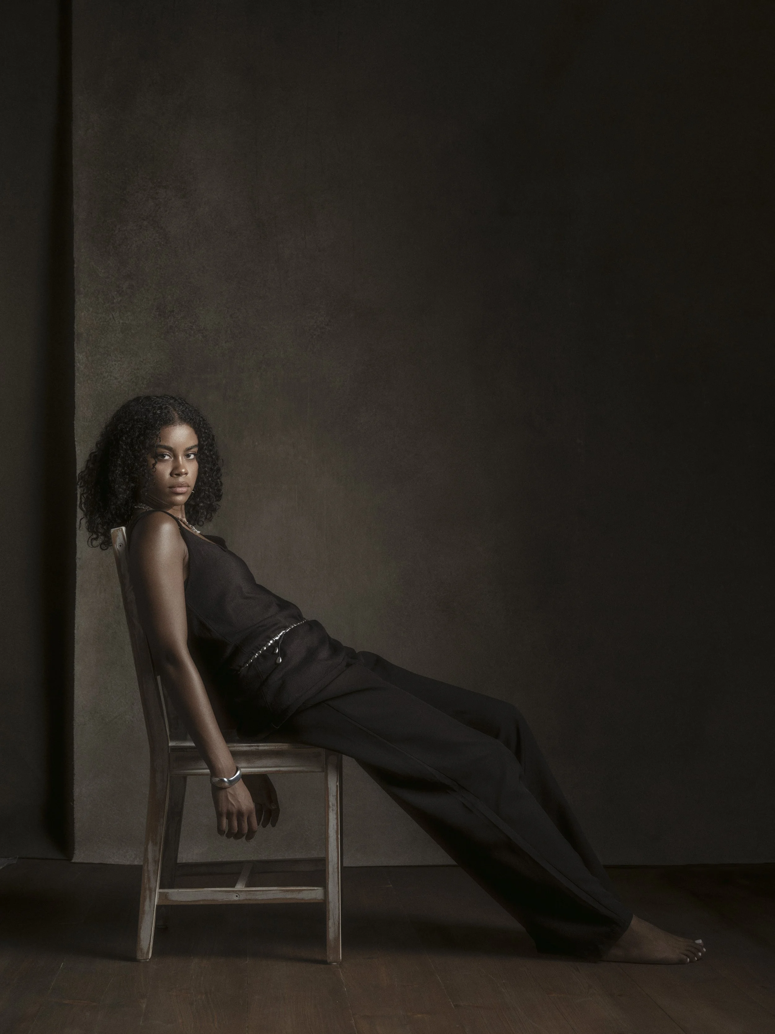 A woman with curly hair and dark clothing sitting on a wooden chair against a dark background, with her left arm hanging down to the floor and her right arm resting on the back of the chair.