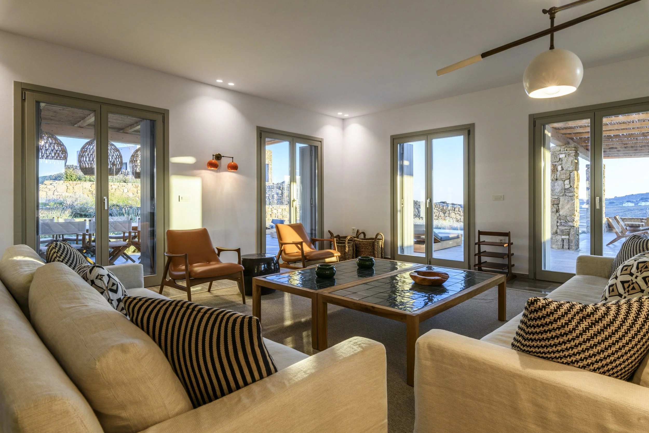 Living room with beige sofas, orange chairs, a wooden coffee table, large windows, and a view of an outdoor patio with stone wall and seating, in a house with modern decor.