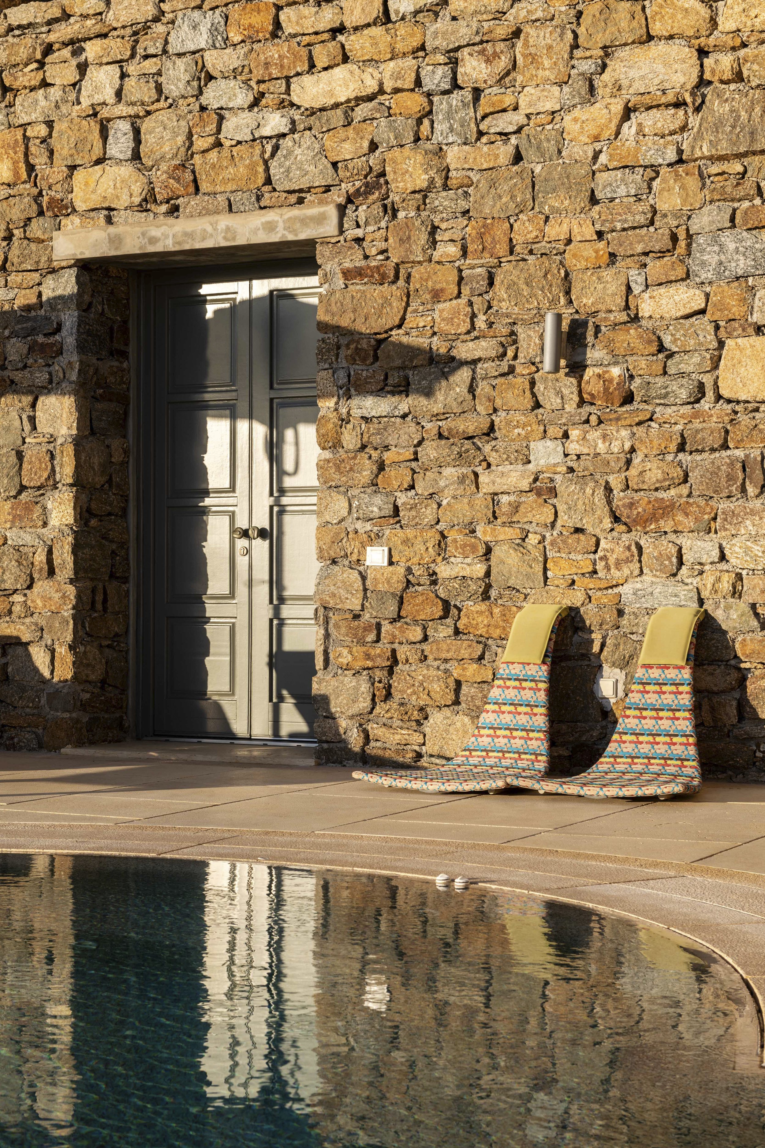 A stone wall with a closed gray door and a metallic outdoor light fixture. Two colorful lounge chairs with patterned fabric are placed next to the wall. In front, a swimming pool reflects the wall and chairs.