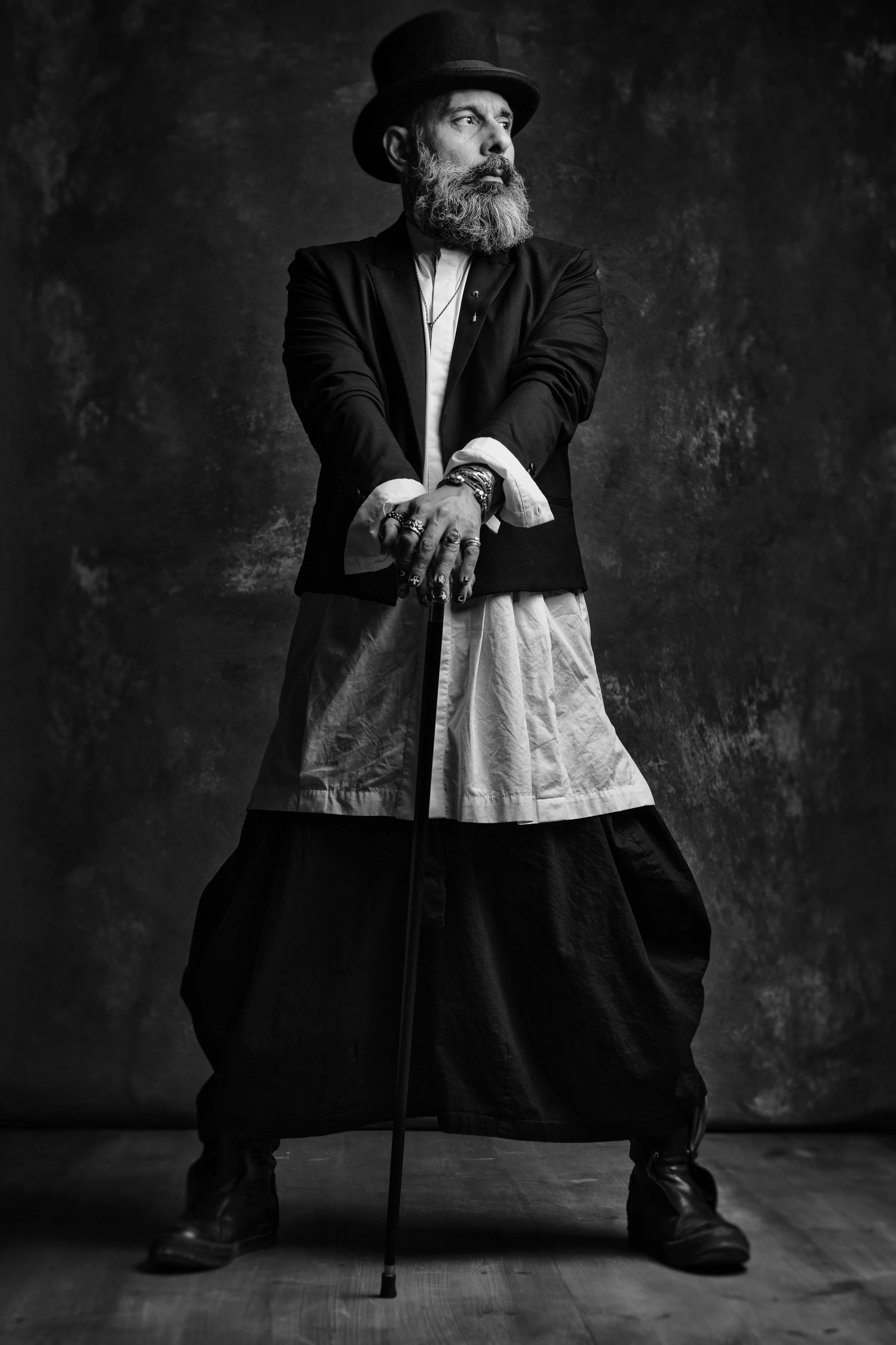 A black and white photo of a man with a beard and mustache, wearing a top hat, a suit with a vest, layered clothing, and outdoor boots. He is holding a cane and has various rings and bracelets. The background is dark and textured.