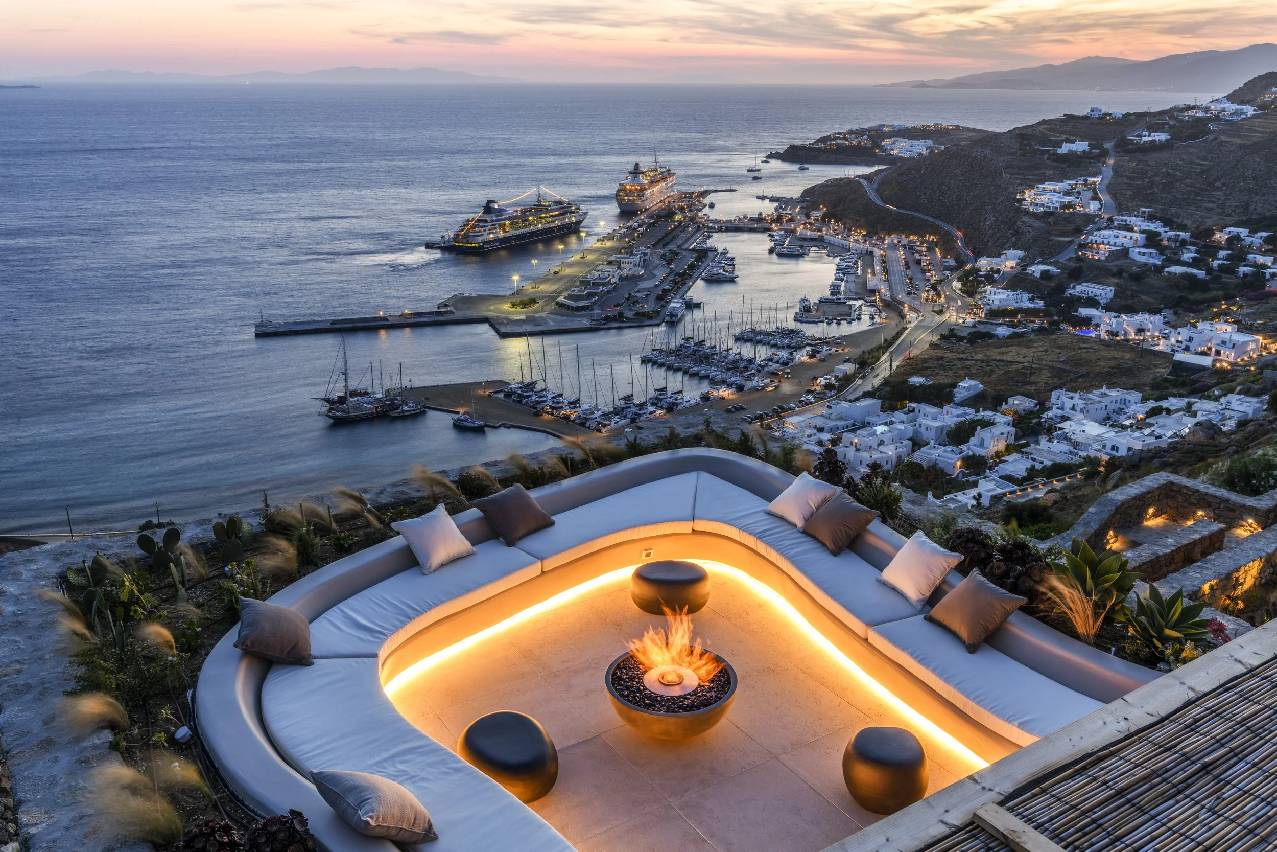 A seaside harbor at sunset with yachts docked and white buildings on the hillside, viewed from a terrace with a circular seating area and a fire pit surrounded by cushions.