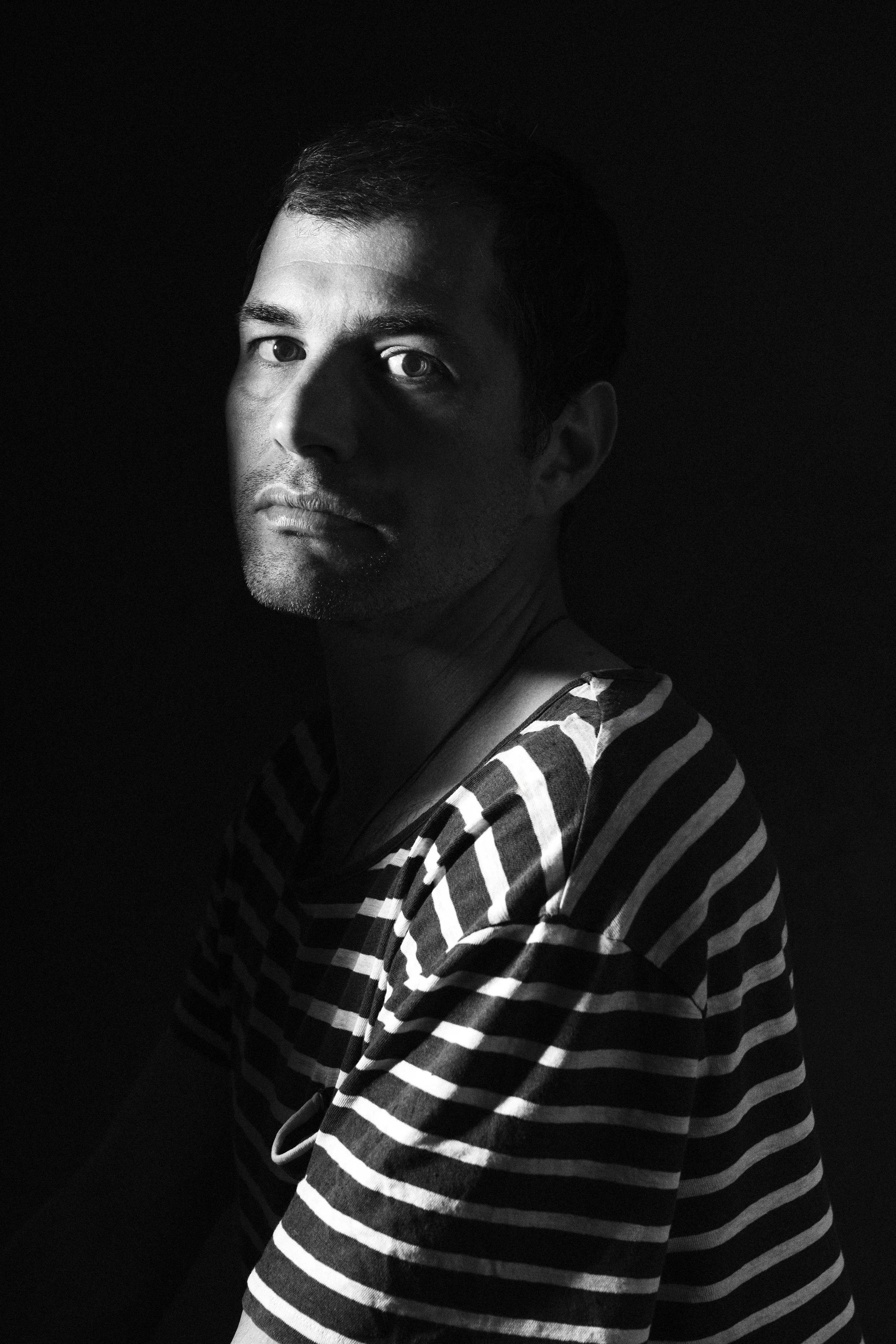 Black and white portrait of a man with short dark hair, wearing a striped shirt, looking at the camera with a serious expression, against a dark background.