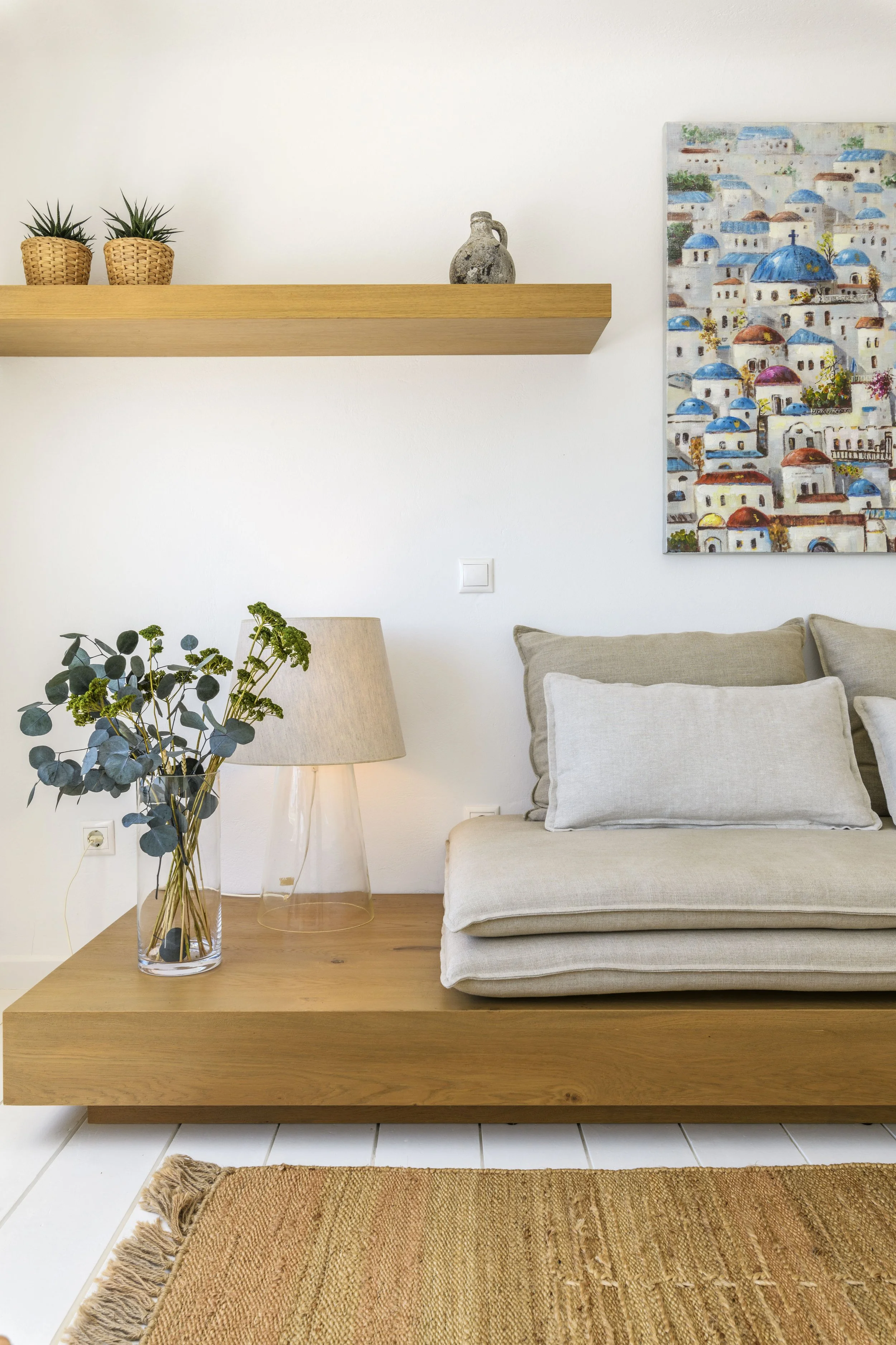 Living room scene with a beige sofa, two cushions, a wooden coffee table, a glass vase with green leafy branches, a table lamp with a beige shade, a wall shelf with two small potted plants, a decorative vase, a colorful painting of a town with white 