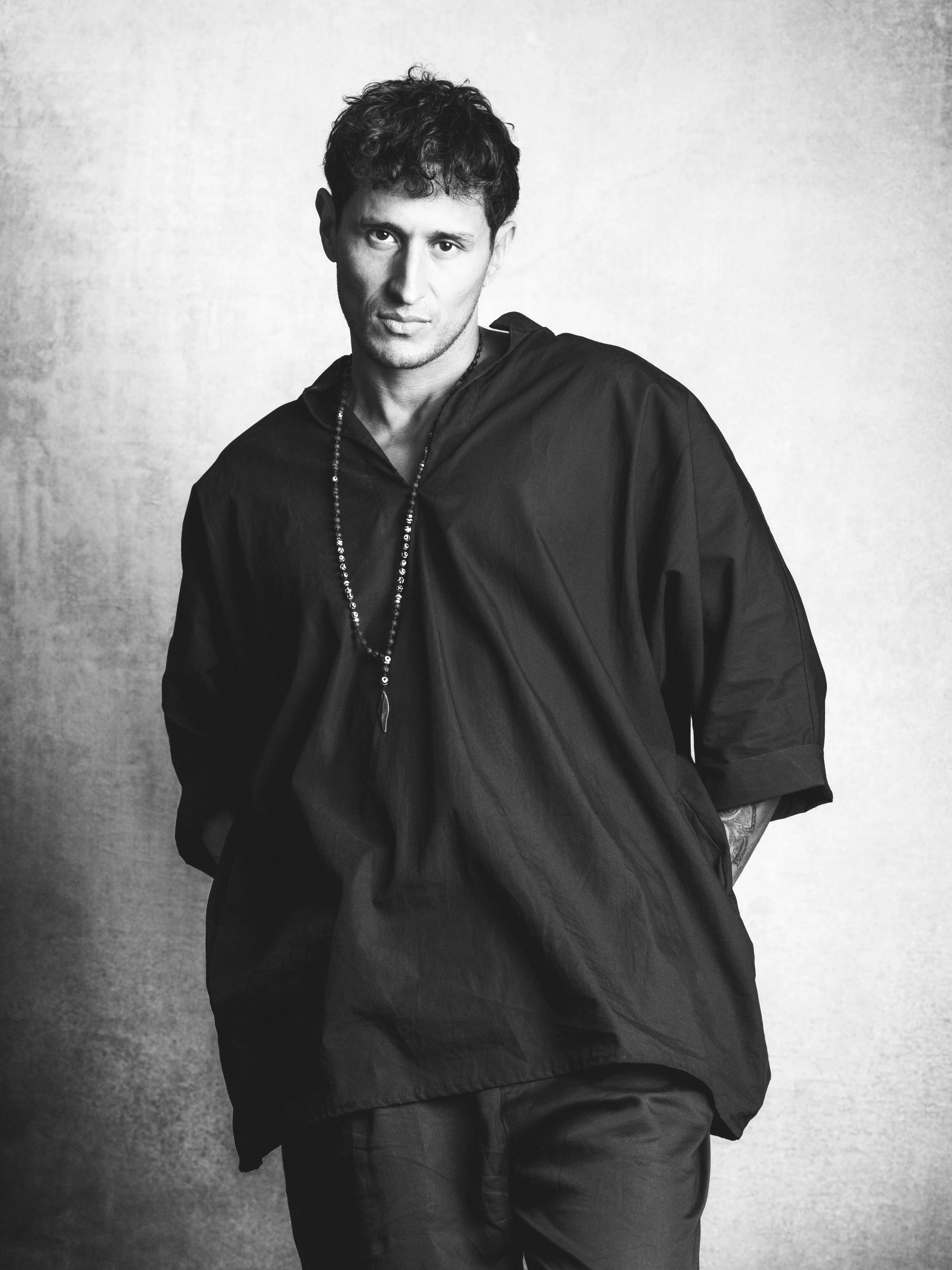 Black and white portrait of a young man with short, curly hair, wearing a dark jacket and a beaded necklace, standing against a plain background.