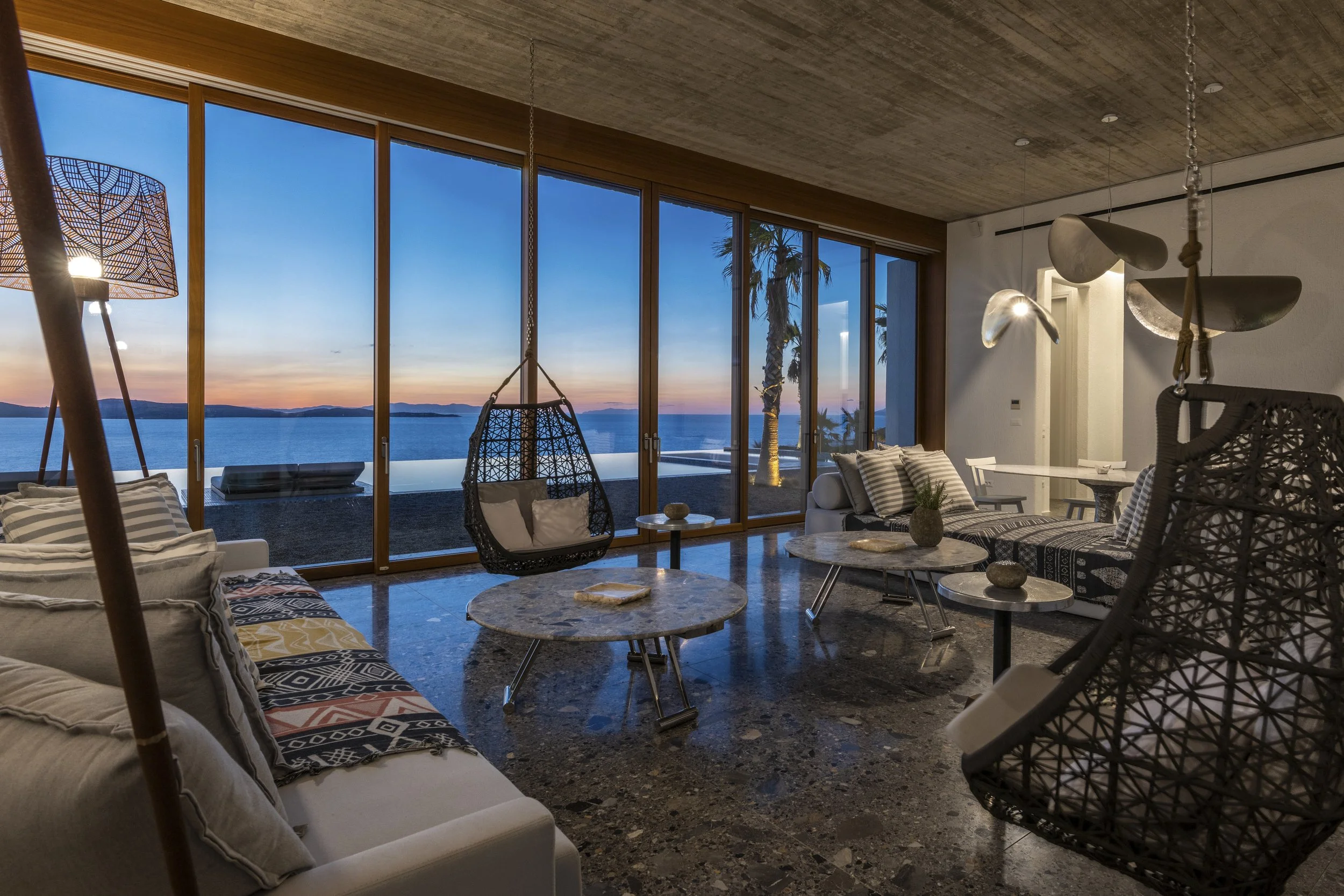 Living room of a luxury villa I Mykonos with large windows showing a sunset view over water and palm trees, decorated with modern and bohemian furniture and lighting.
