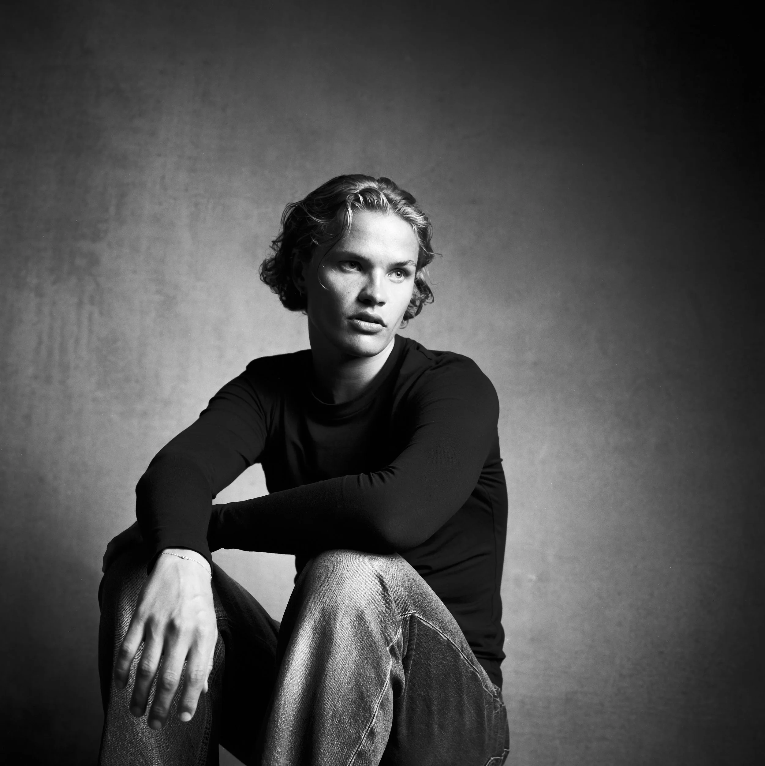 Black and white photo of a woman with curly hair looking upward, with her right hand resting on her chin and her left arm crossing her body, wearing a long-sleeve top and jeans, against a plain dark background.