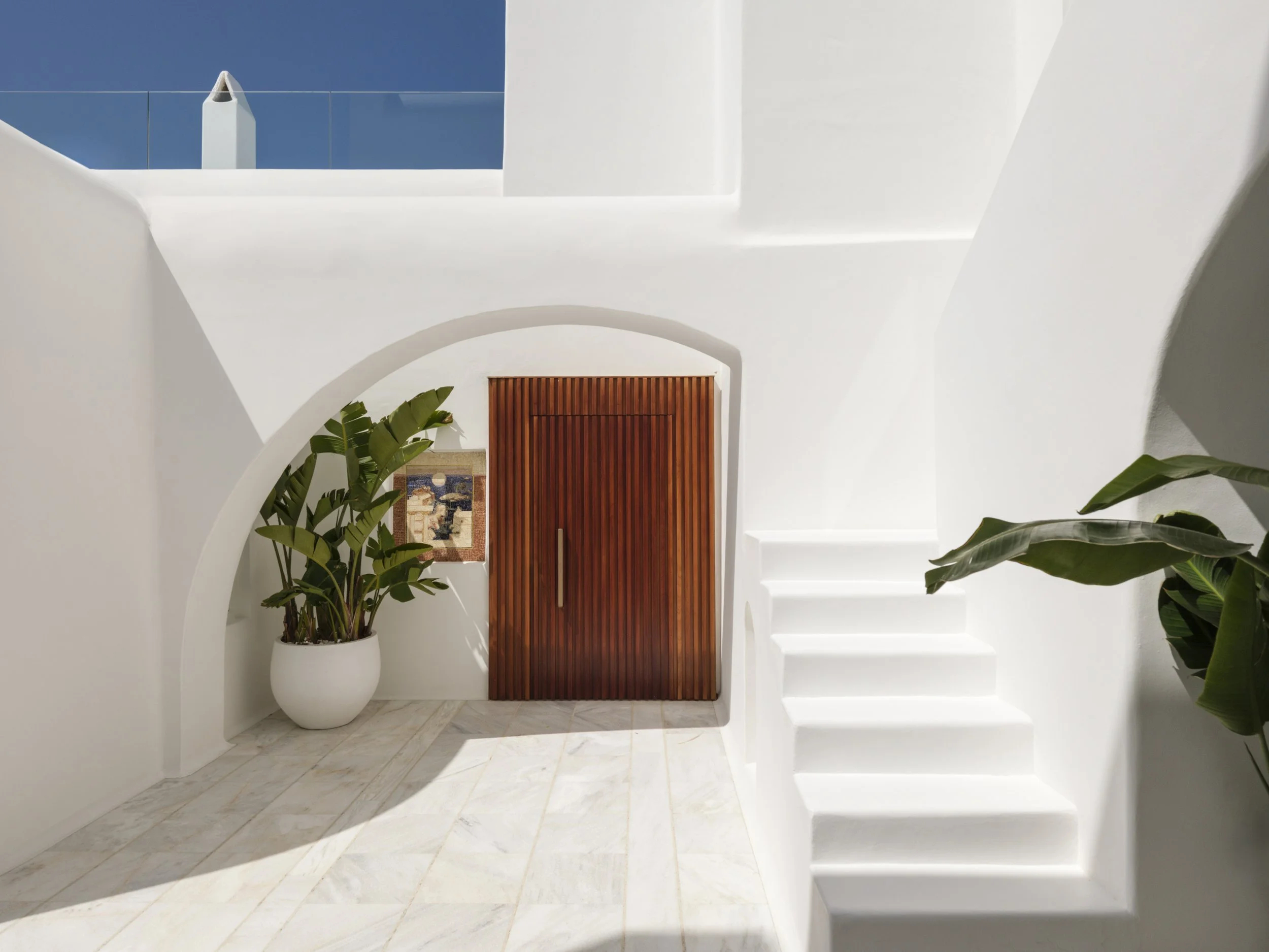 A white staircase with potted plants on either side leading to a wooden door beneath an arched ceiling. Bright natural light casts shadows on the white walls and marble floor.