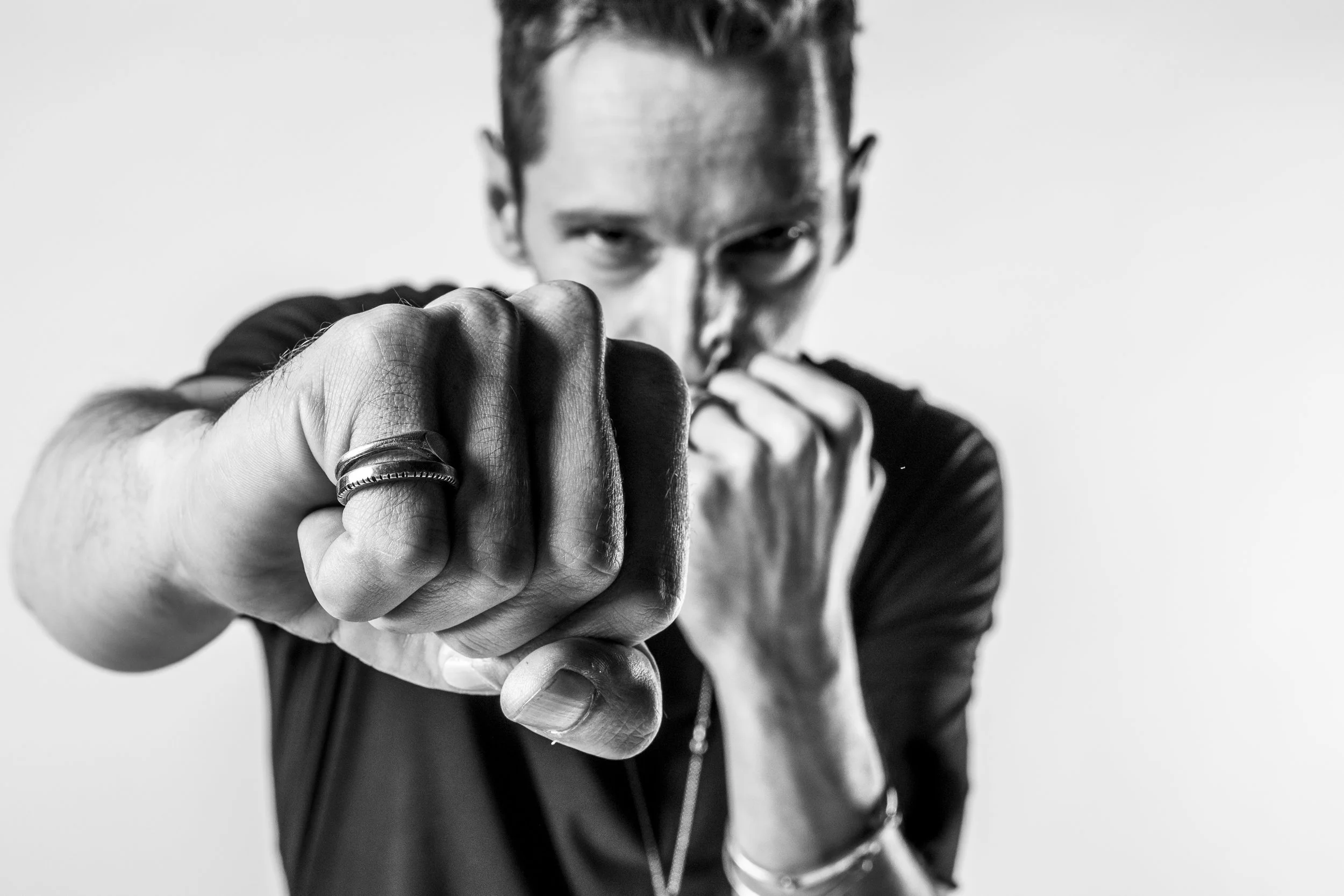 A man with a serious expression, wearing a black t-shirt, pointing directly at the camera, with his hand and fist prominently in focus, in black and white.