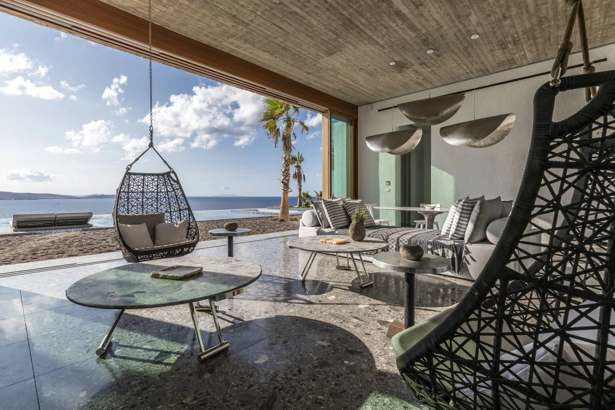 Modern living space of a luxury Villa located in Mykonos island with large glass doors opening to a view of the Aegean Sea , palm trees, and blue sky. Outdoor seating includes woven hanging chair and white sofa with striped cushions, with marble-topp