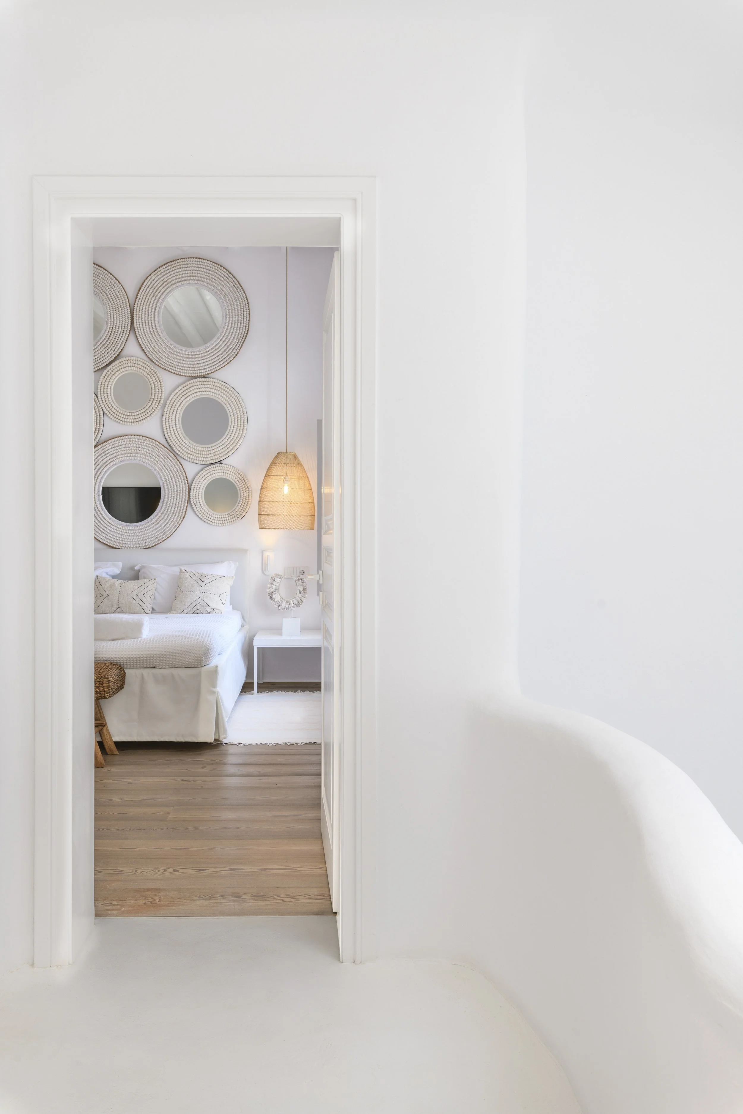 Bright bedroom interior of a luxury villa on Mykonos island, viewed through a white doorway, showcasing chic bohemian decor with round woven wall mirrors and a wicker pendant light.