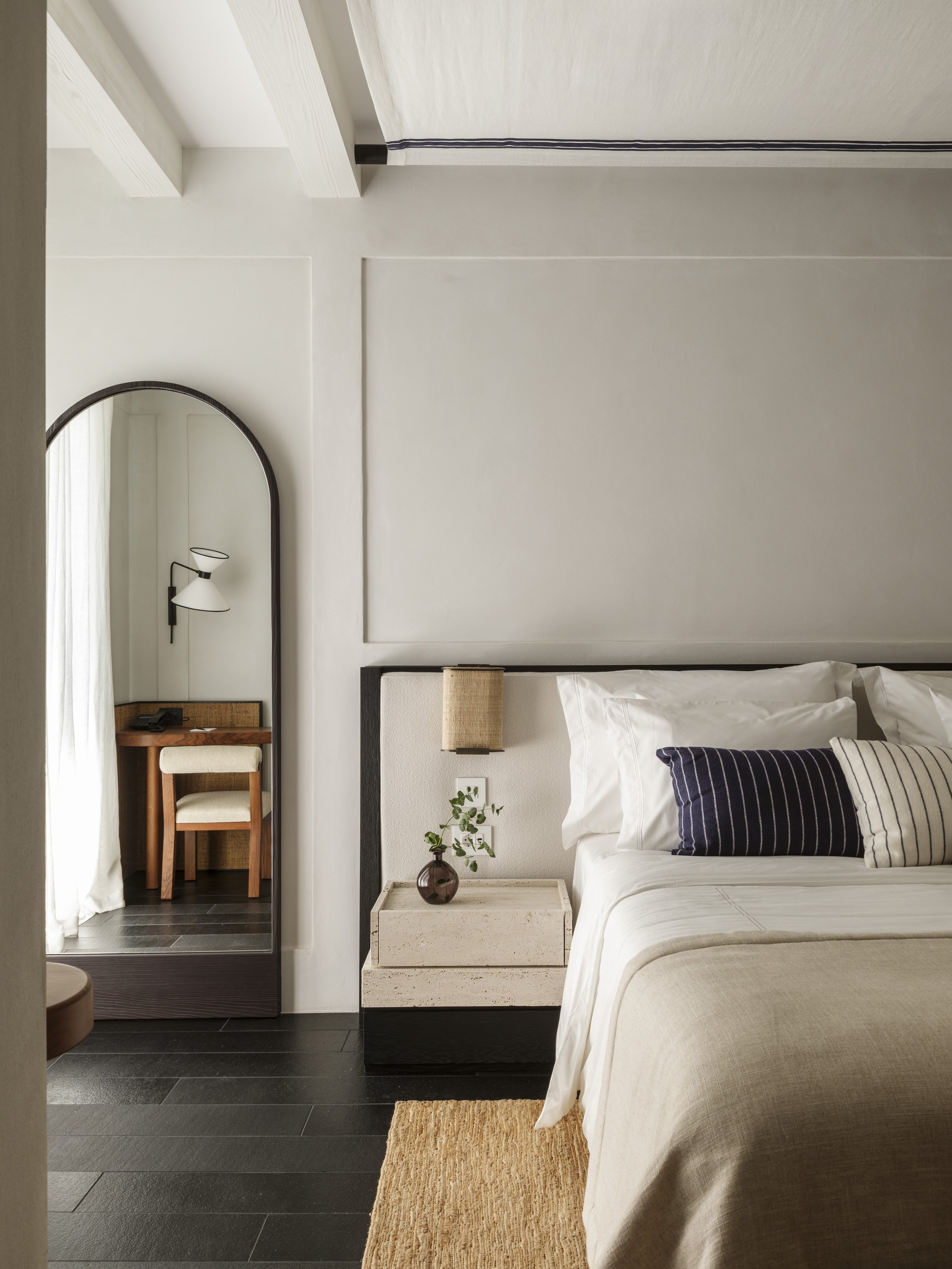A modern bedroom with a bed, striped pillow, bedside table with a small plant, and a mirror with a seat and a lamp in the reflection.