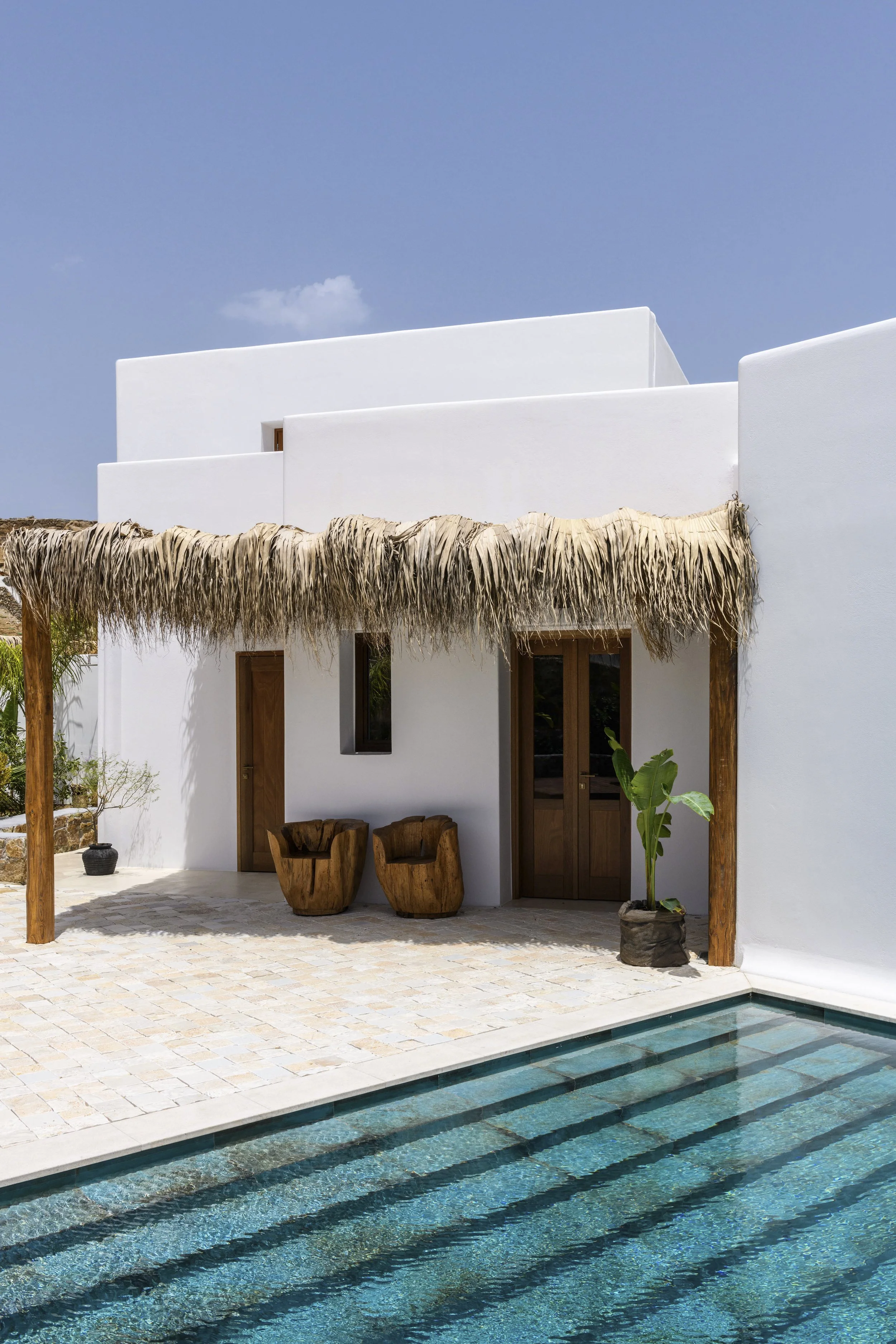White modern building with thatched roof accents, wooden chairs, potted plants, and a swimming pool with steps in a sunny outdoor setting.