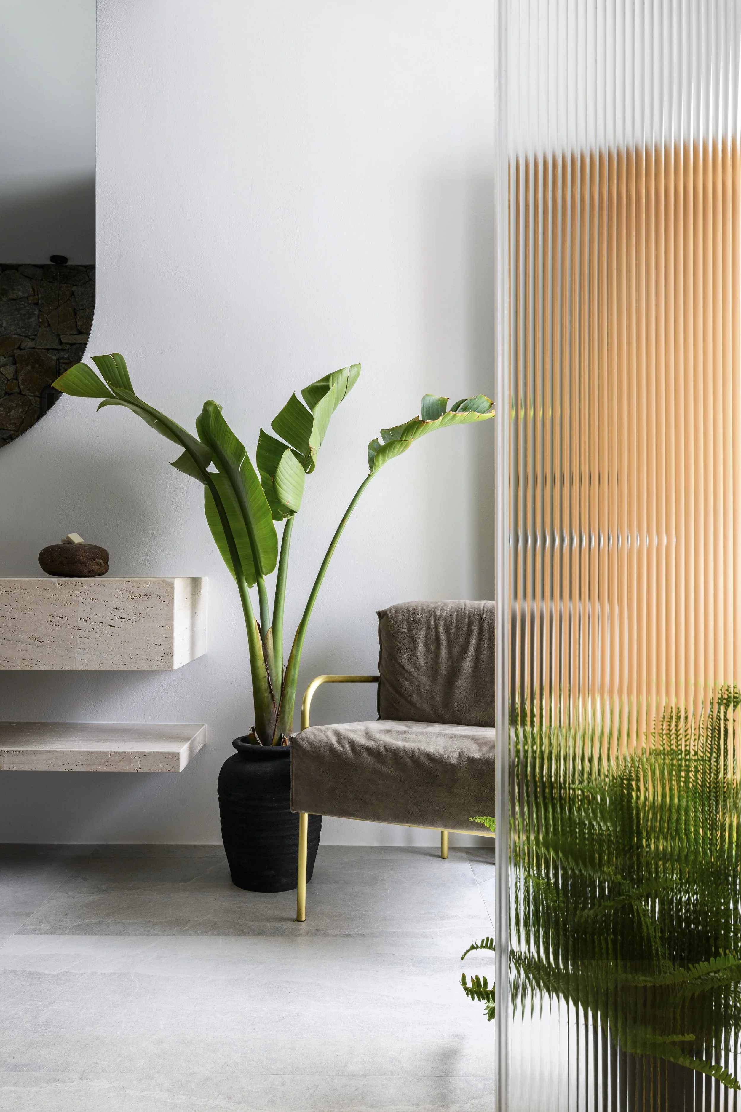 A modern interior with a large green potted plant, beige armchair with gold legs, a floating shelf with decorative items, and a partial view of a room with stone wall, all behind a stylish, semi-transparent wooden slat room divider with green plants at the bottom.