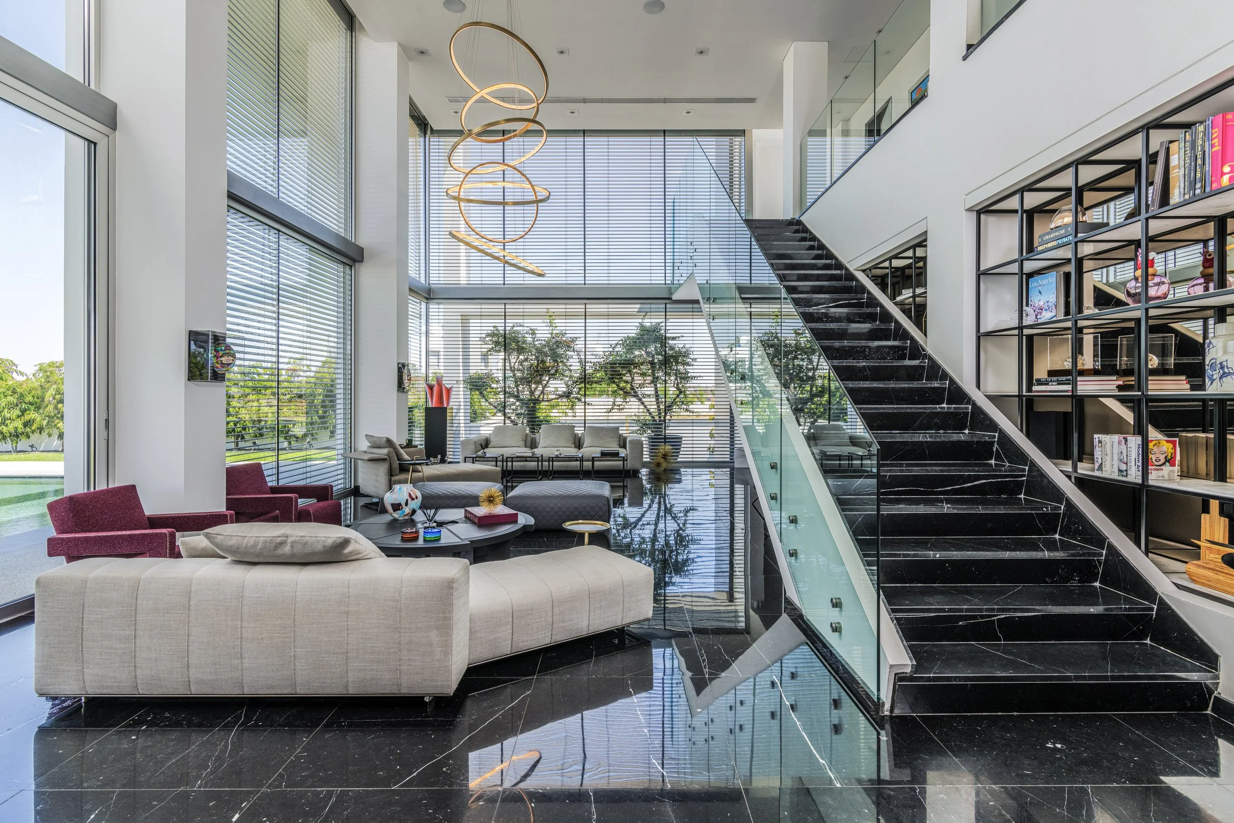 Modern interior of a spacious lobby with large windows, a black marble staircase with glass railing, a bookshelf filled with books and decorative items, stylish seating including beige and red chairs, and a spiral pendant light fixture hanging from t