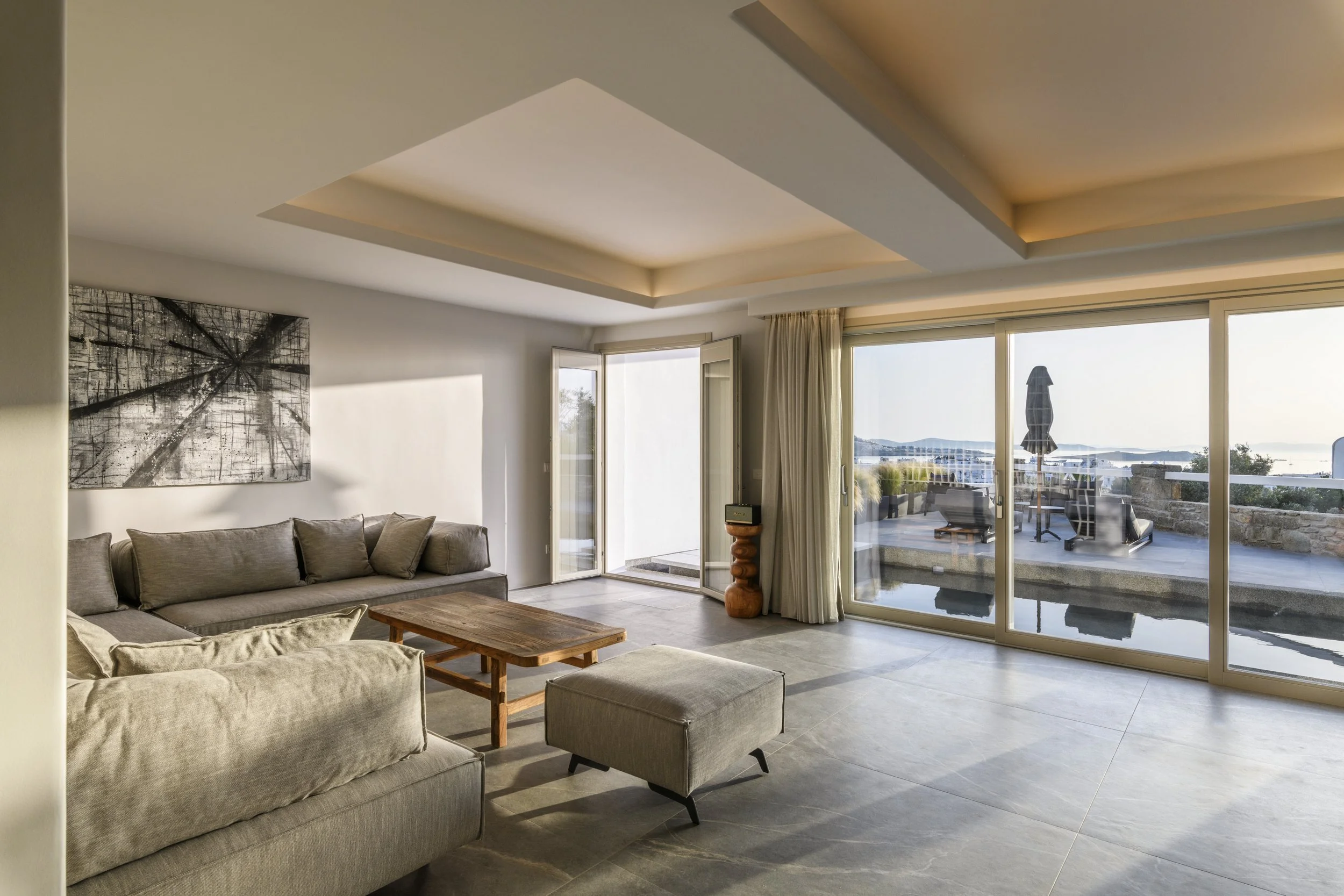Living room with beige sofas, a wooden coffee table, a large abstract black and white wall art, and large glass doors with curtains leading to an outdoor patio with a view of water and islands.