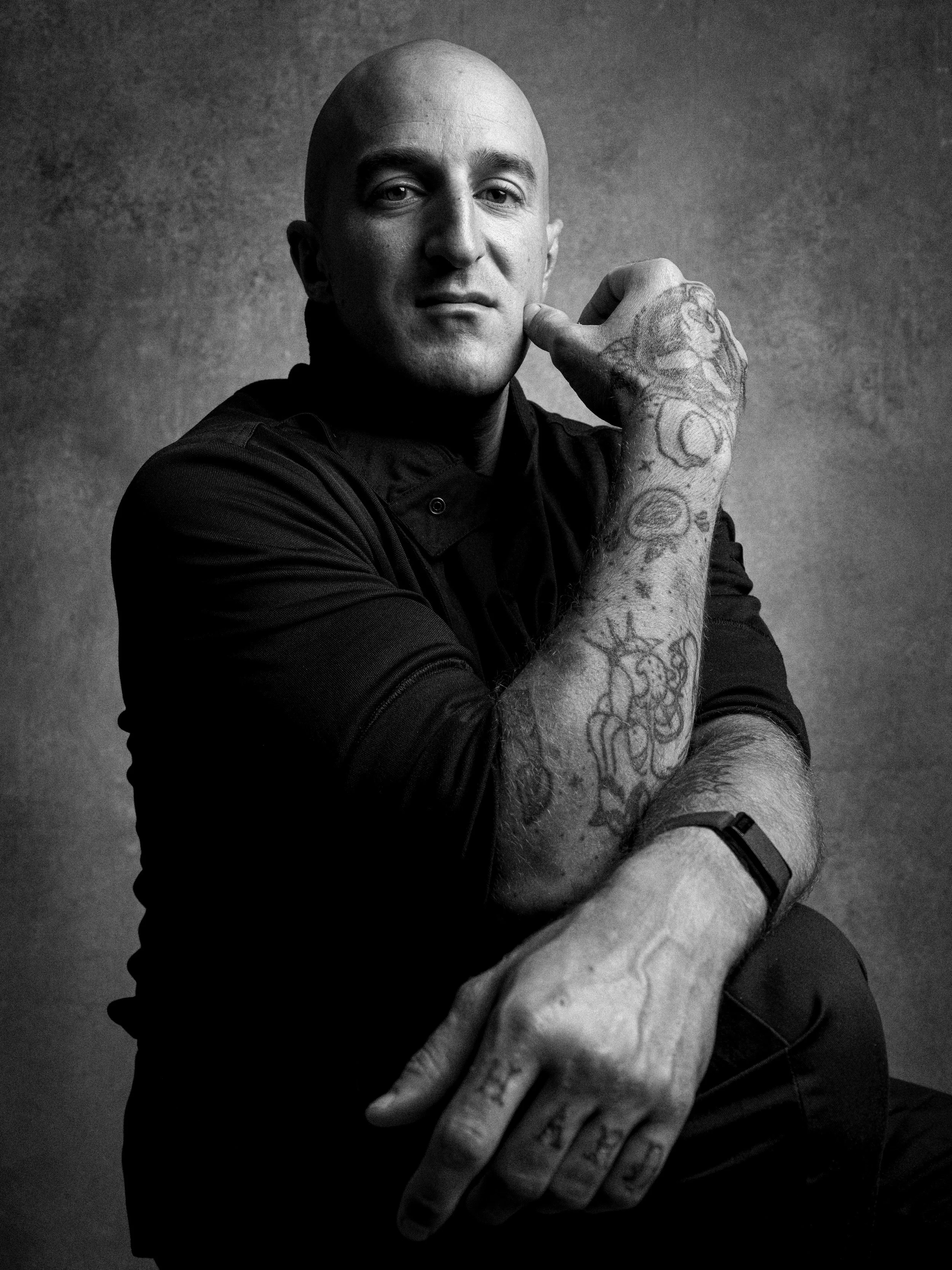 A black and white portrait of a bald man with tattoos on his arm, looking at the camera with a confident expression, resting his chin on his hand.