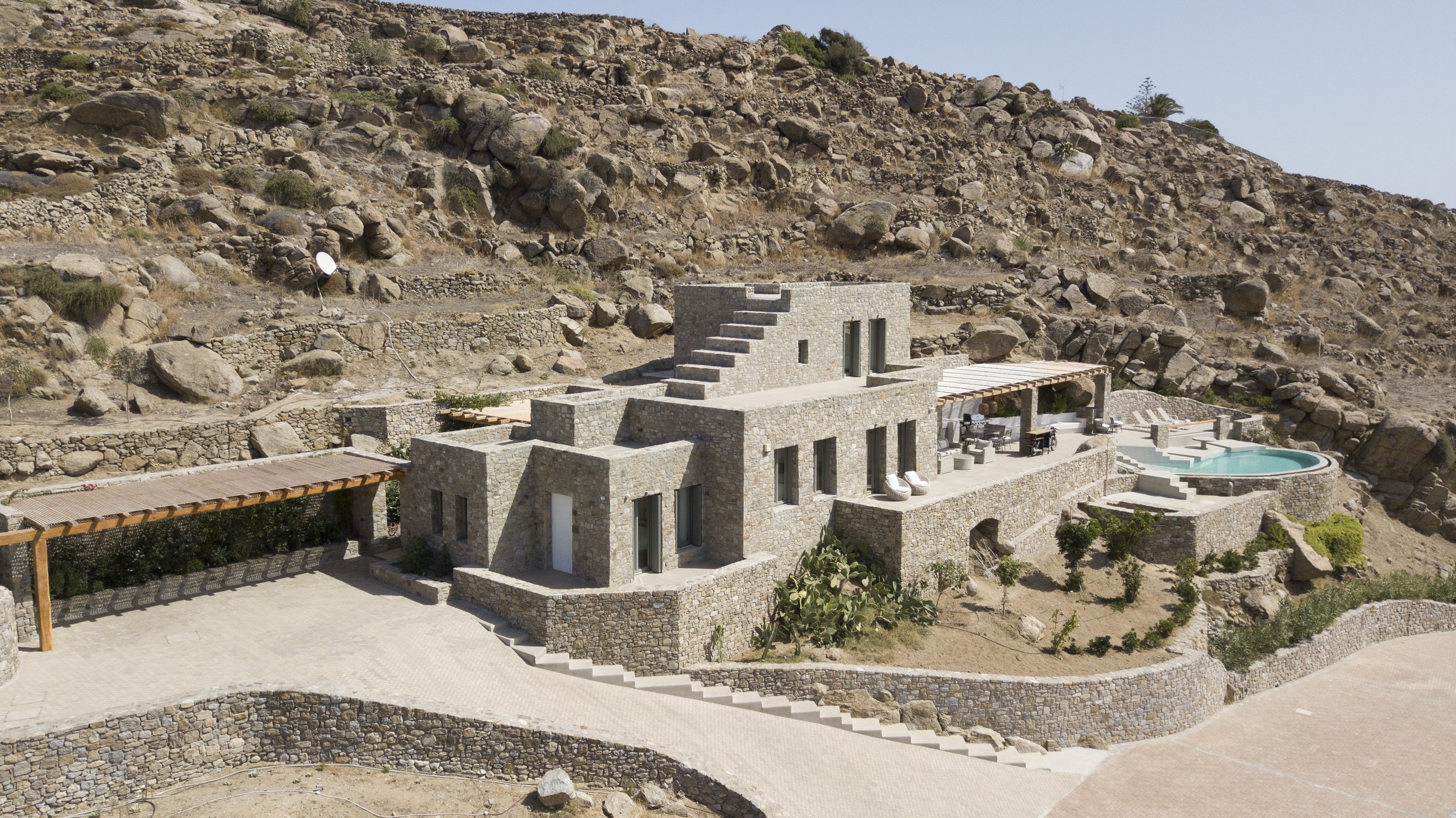 Modern house built into a rocky hillside with stone exterior walls, outdoor seating area, swimming pool, and desert landscape surrounding it.