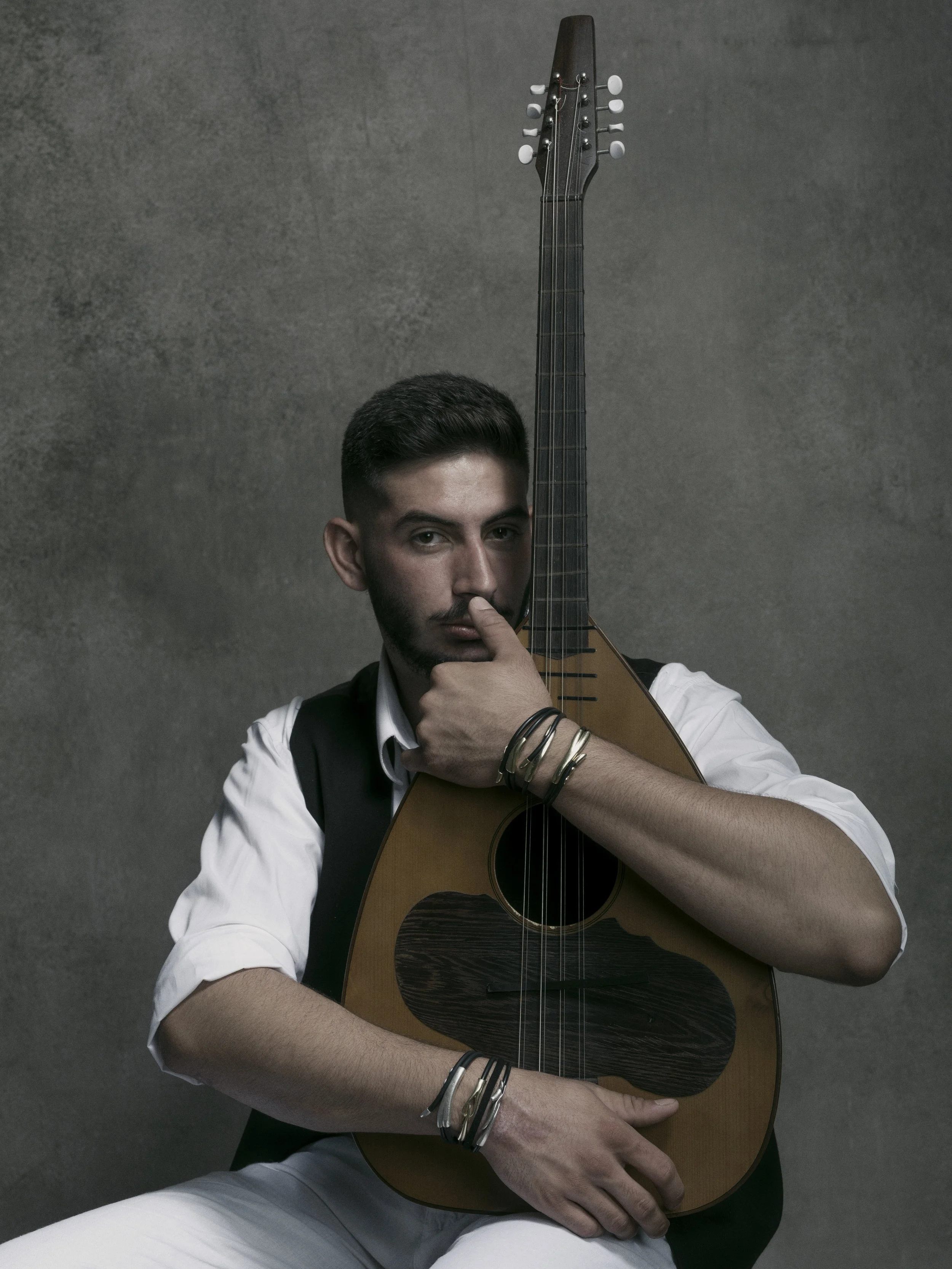 A young man with short dark hair and a beard, wearing a white shirt and black vest, holds an acoustic guitar vertically with one hand resting on the body and the other on the neck, against a plain gray textured wall background.