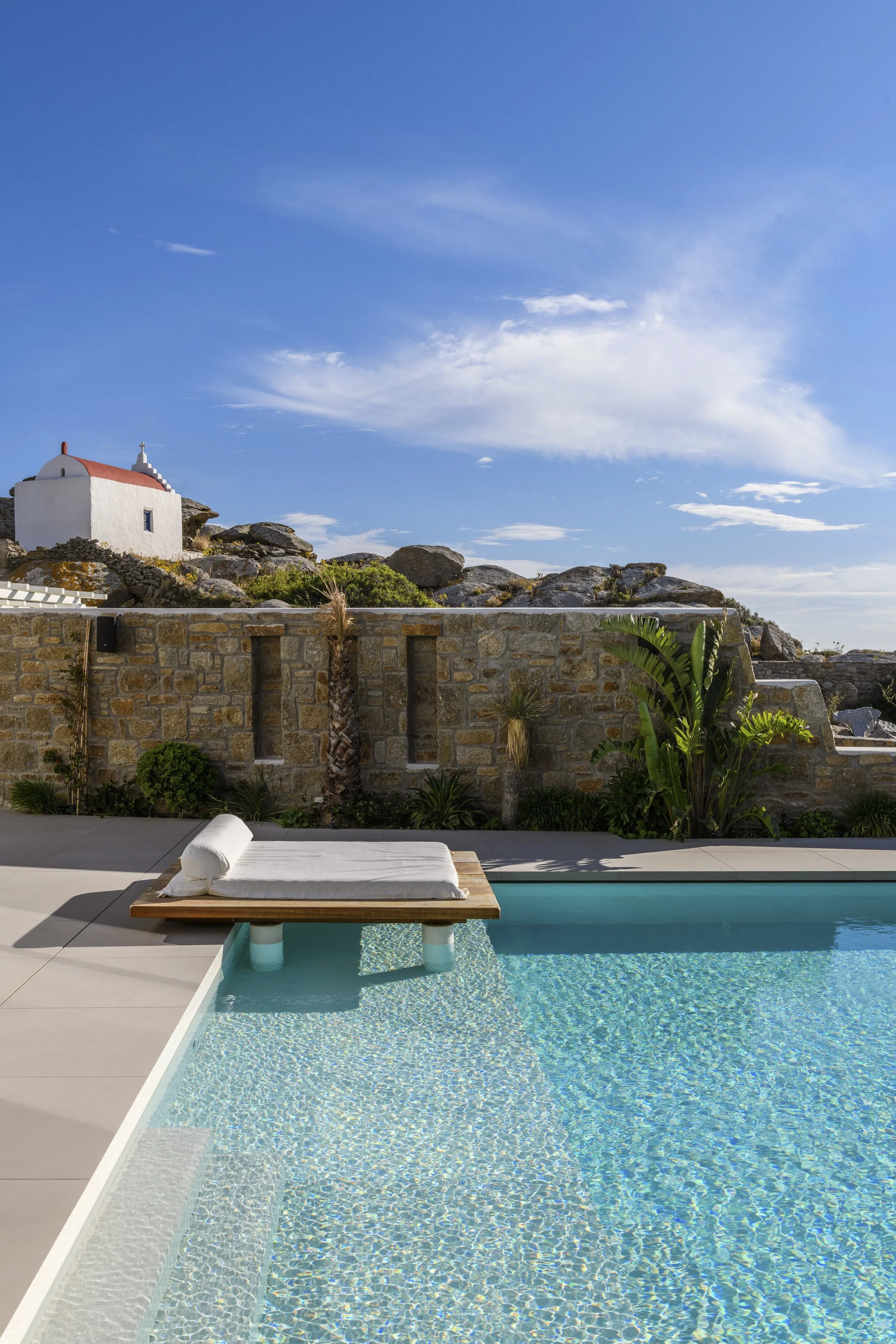 A swimming pool with a wooden lounge chair and cushion in the foreground, stone wall and bushes beyond, a white building with a red roof on rocks to the left, a blue sky with scattered clouds overhead.