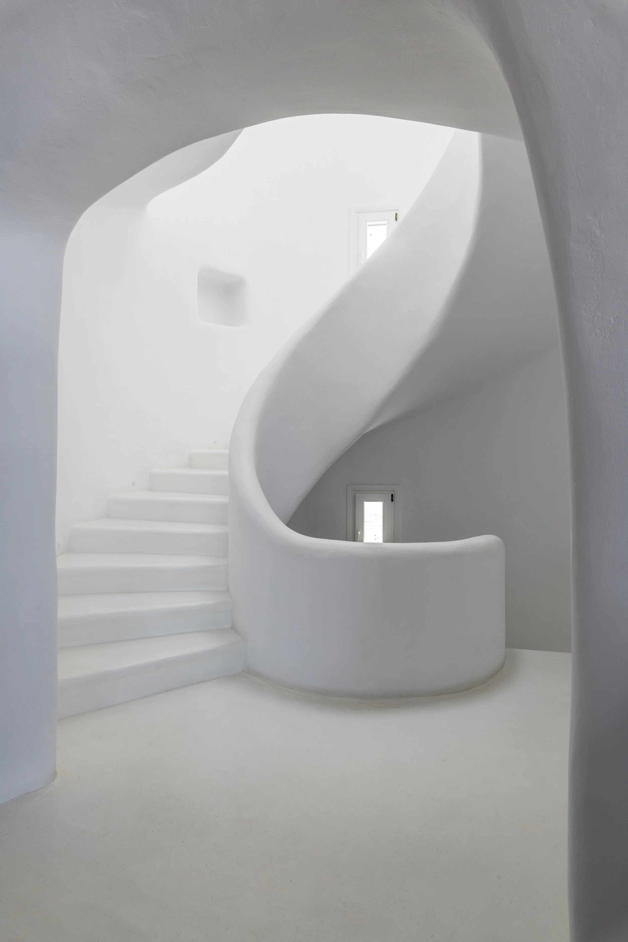 Modern luxury villa interior on Mykonos island featuring a smooth, minimalist white concrete spiral staircase with organic, flowing architectural lines.