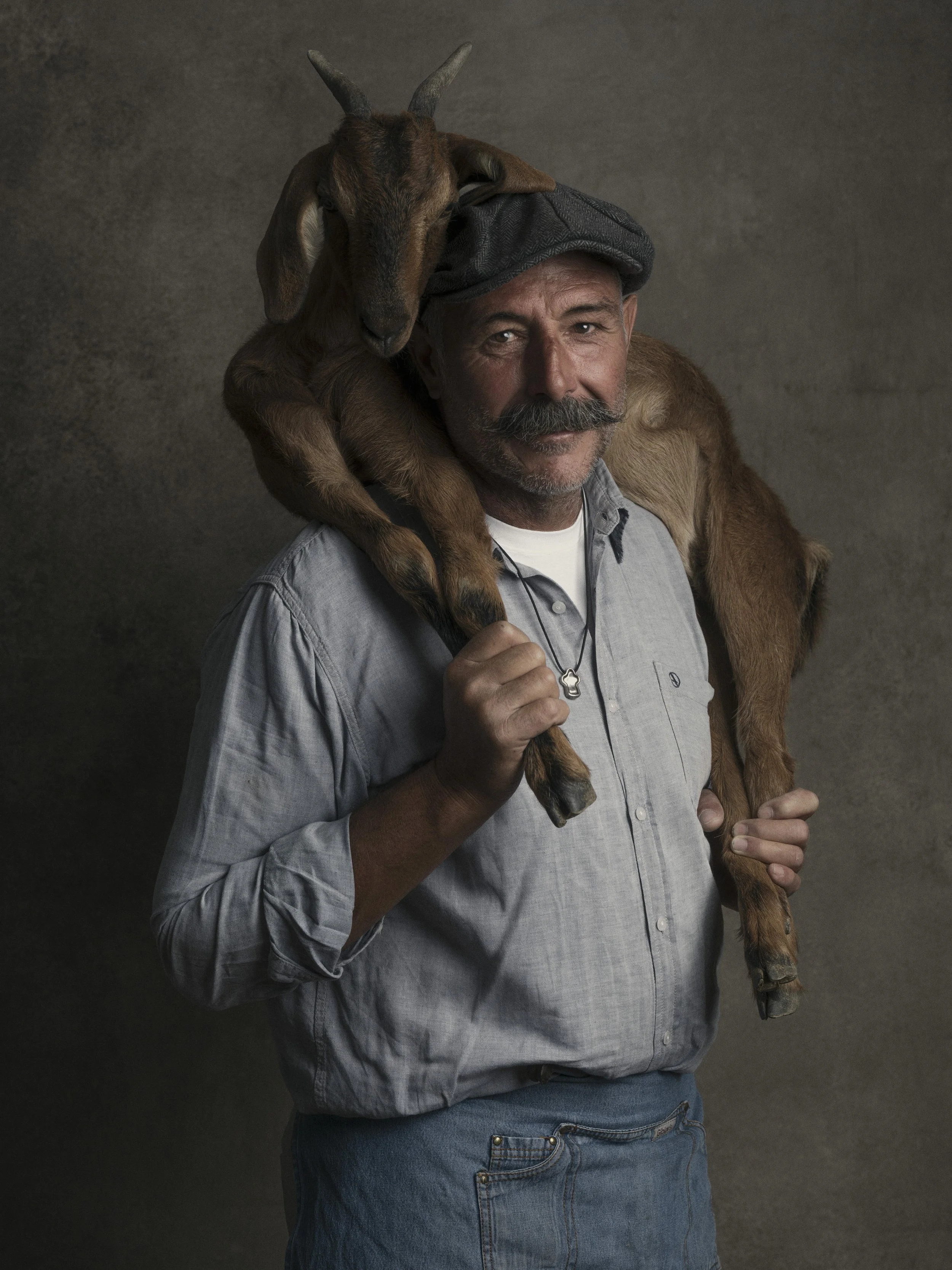 A man with a gray shirt, blue jeans, a gray cap, and a mustache holding a young goat on his shoulder against a dark background.