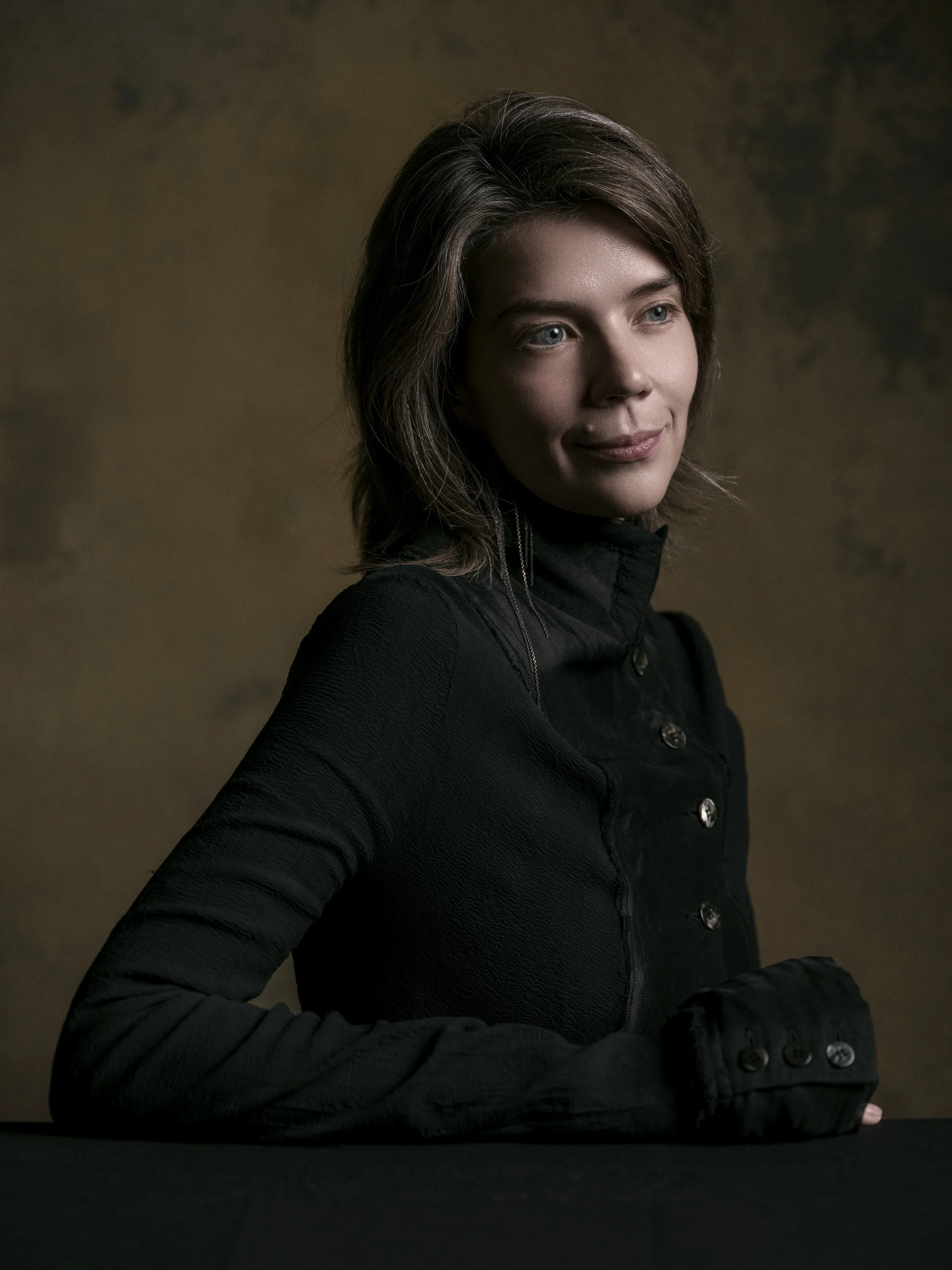 A woman with shoulder-length brown hair, wearing a black jacket, is sitting with her arms crossed on a dark surface against a plain, dark background. She is looking slightly to her right with a slight smile.
