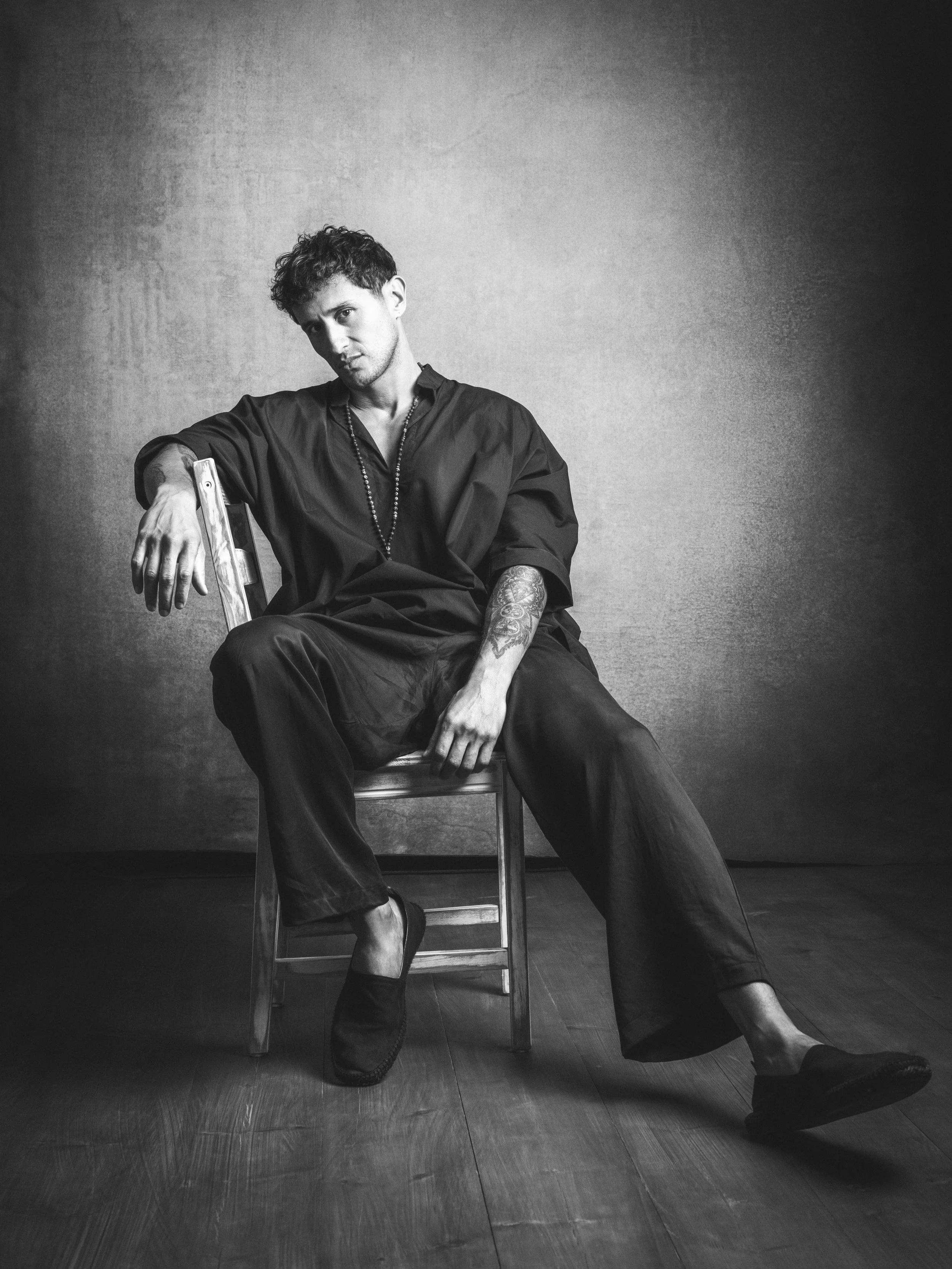 Black and white photo of a young man with curly hair sitting on a chair against a plain background. He is dressed in casual dark clothing with a necklace, has tattoos on his left arm, and is wearing boots. His pose is relaxed, with one arm resting on