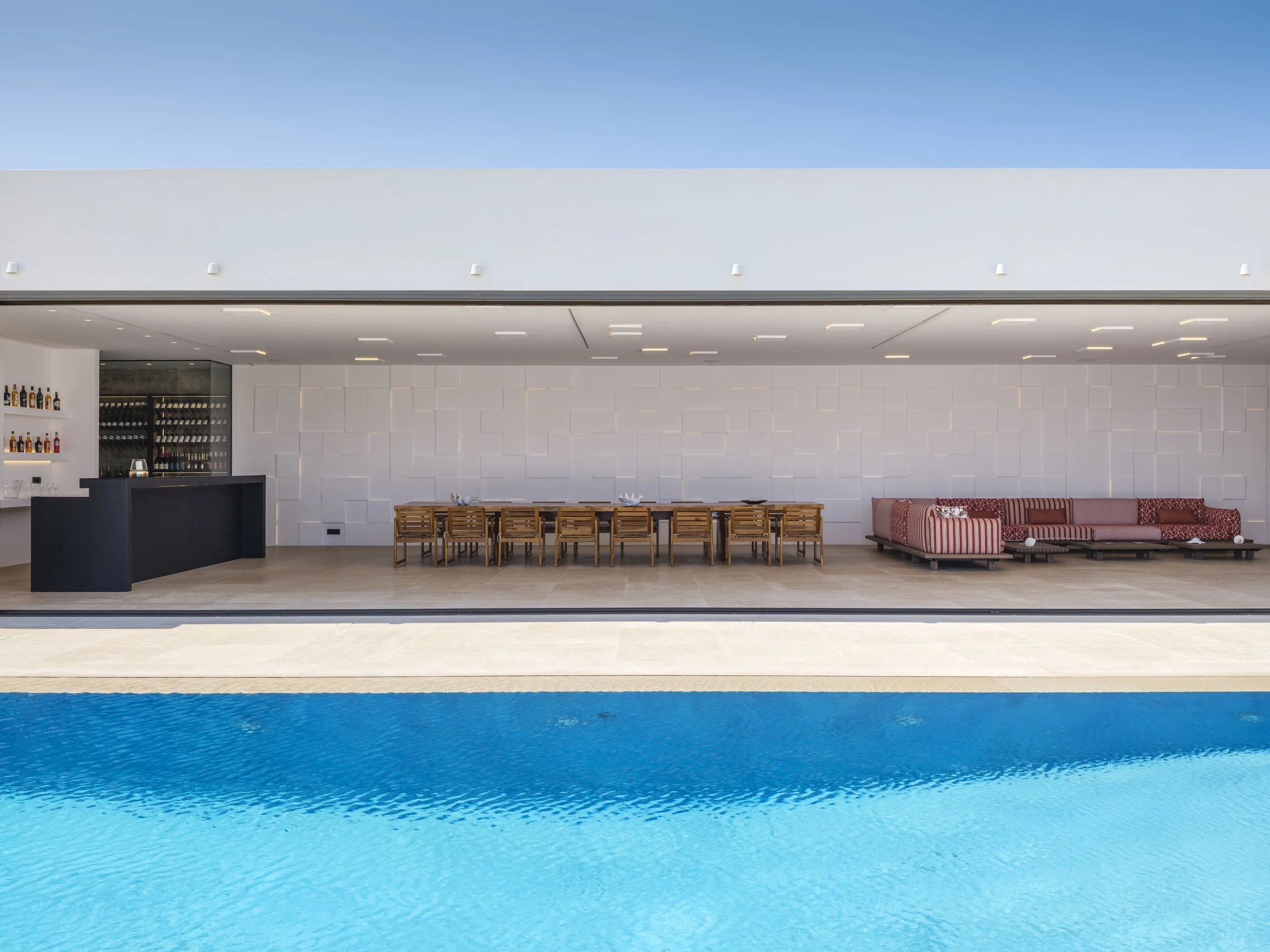 Modern outdoor pool area with a blue swimming pool in the foreground, and a white building with a dining area and wine storage in the background under a clear blue sky.