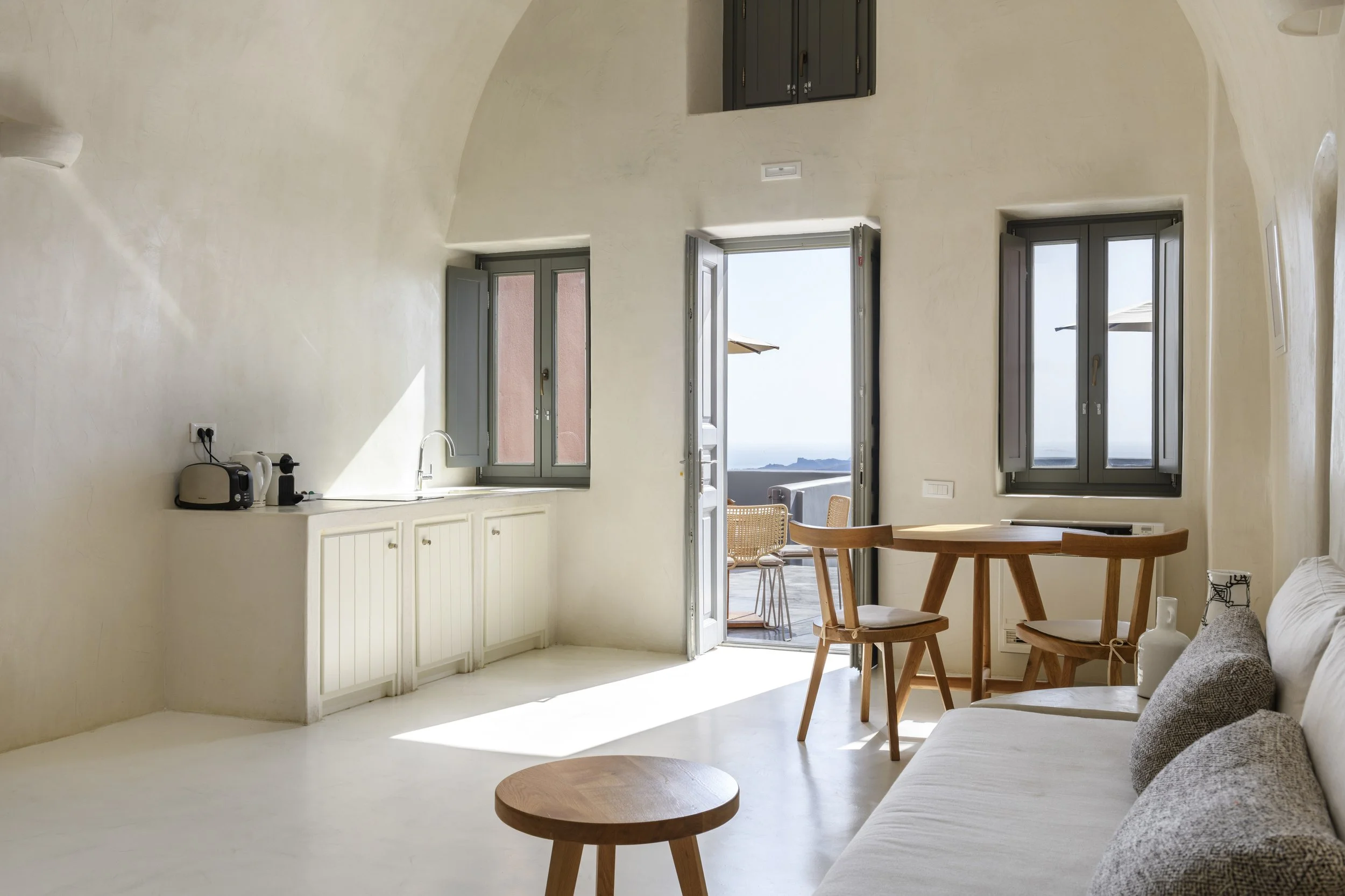 Interior of a bright, minimalist living space with open windows and balcony doors leading to an outdoor view, featuring a wood dining table, kitchen with small appliances, and a sofa.