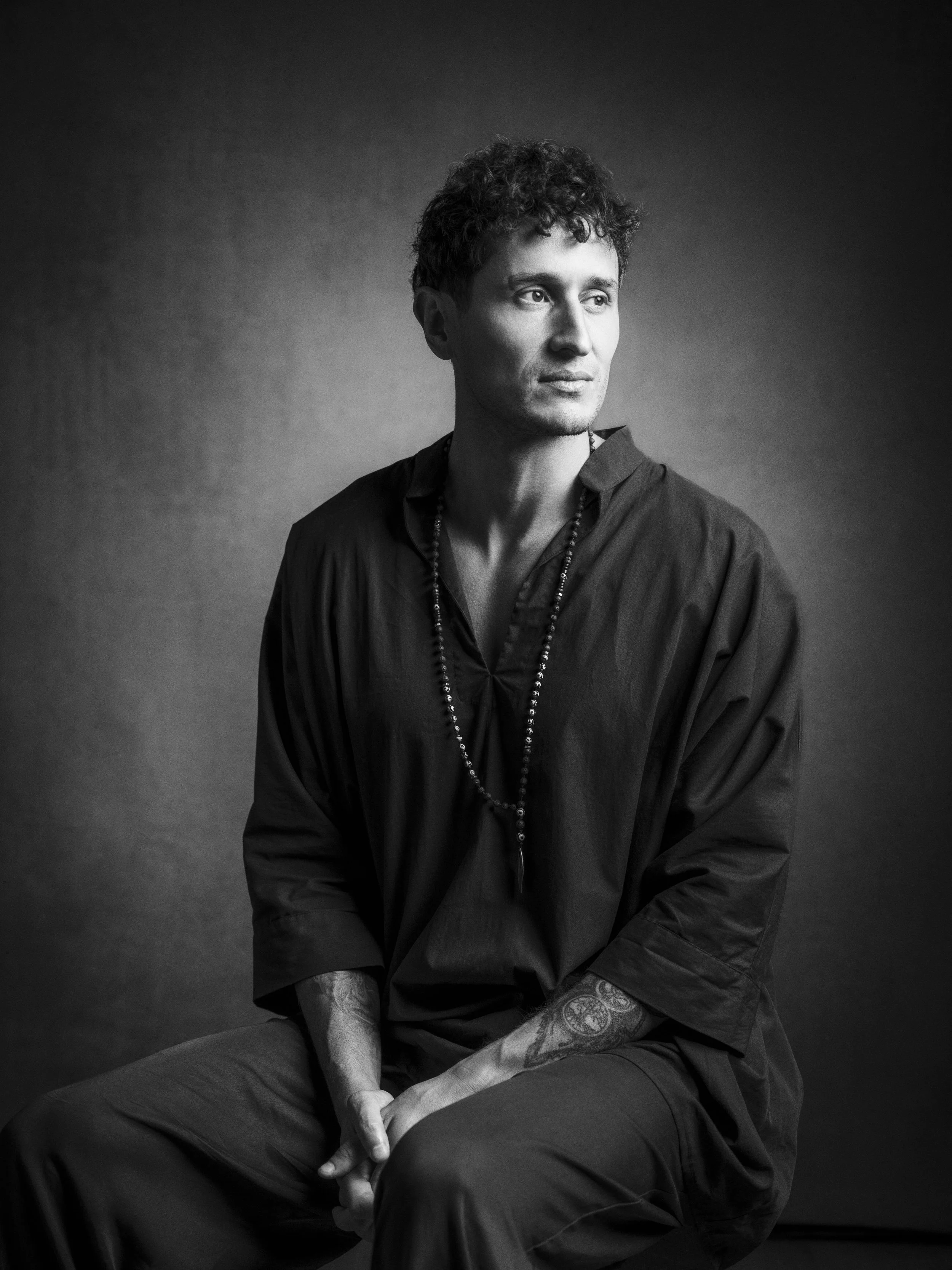 A young man with curly hair and tattoos on his arms, sitting with his hands clasped, looking to his right, wearing a dark long-sleeve shirt and a necklace, against a dark backdrop in a black and white photo.