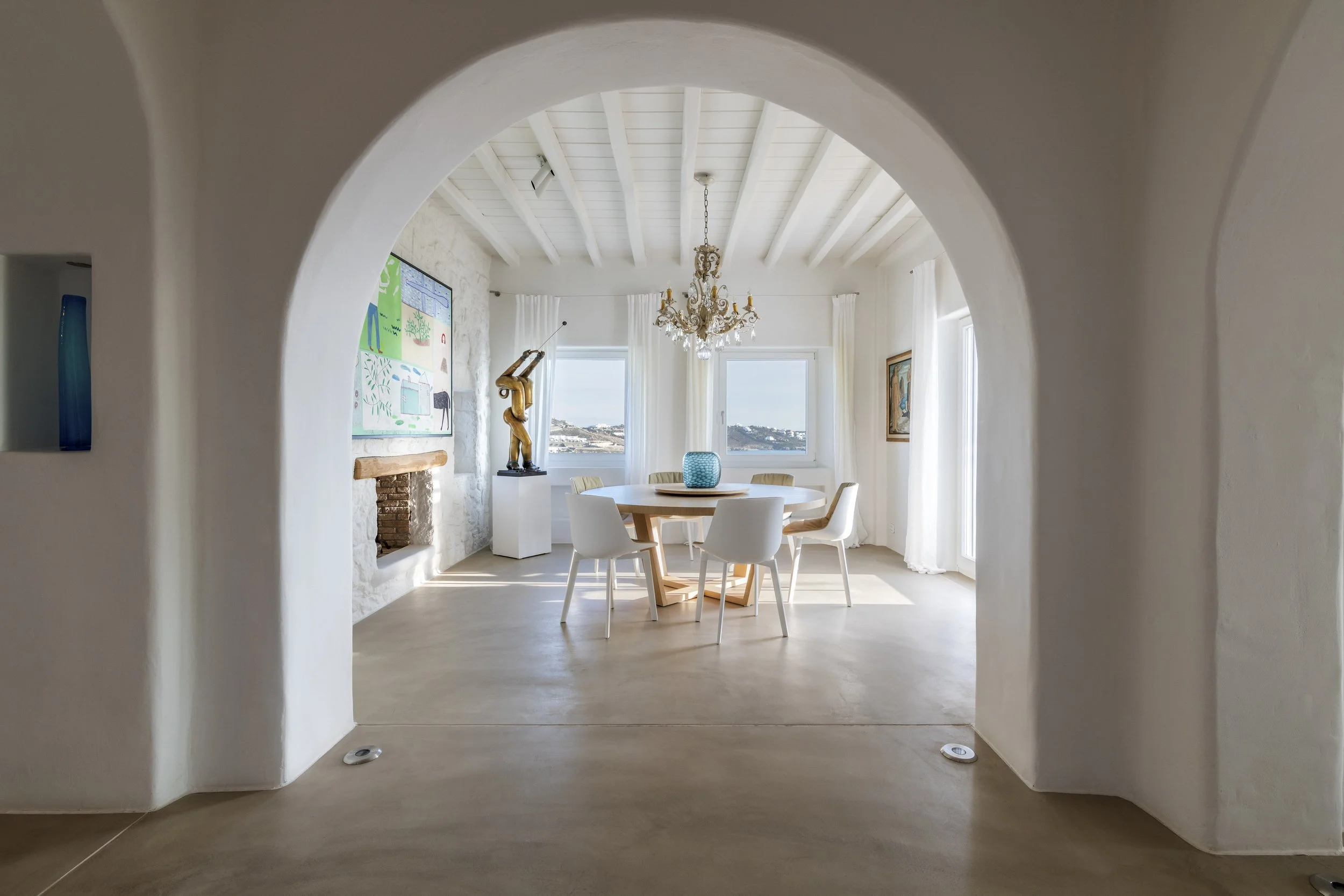 A bright dining room viewed through an archway, featuring a round table with white chairs, a decorative chandelier, and large windows with white curtains, overlooking an outdoor view.