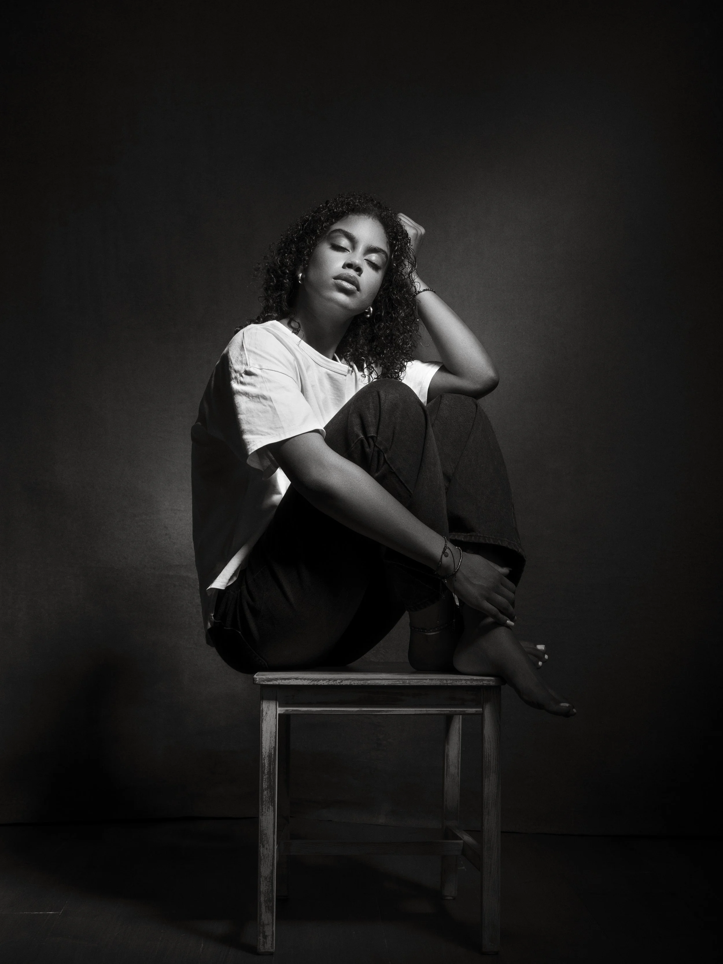 Black-and-white photo of a woman sitting on a wooden table in a confident pose with one leg bent and her arm resting on her knee, wearing a white t-shirt, black pants, and hoop earrings, with her eyes closed and hair styled in natural curly locks.