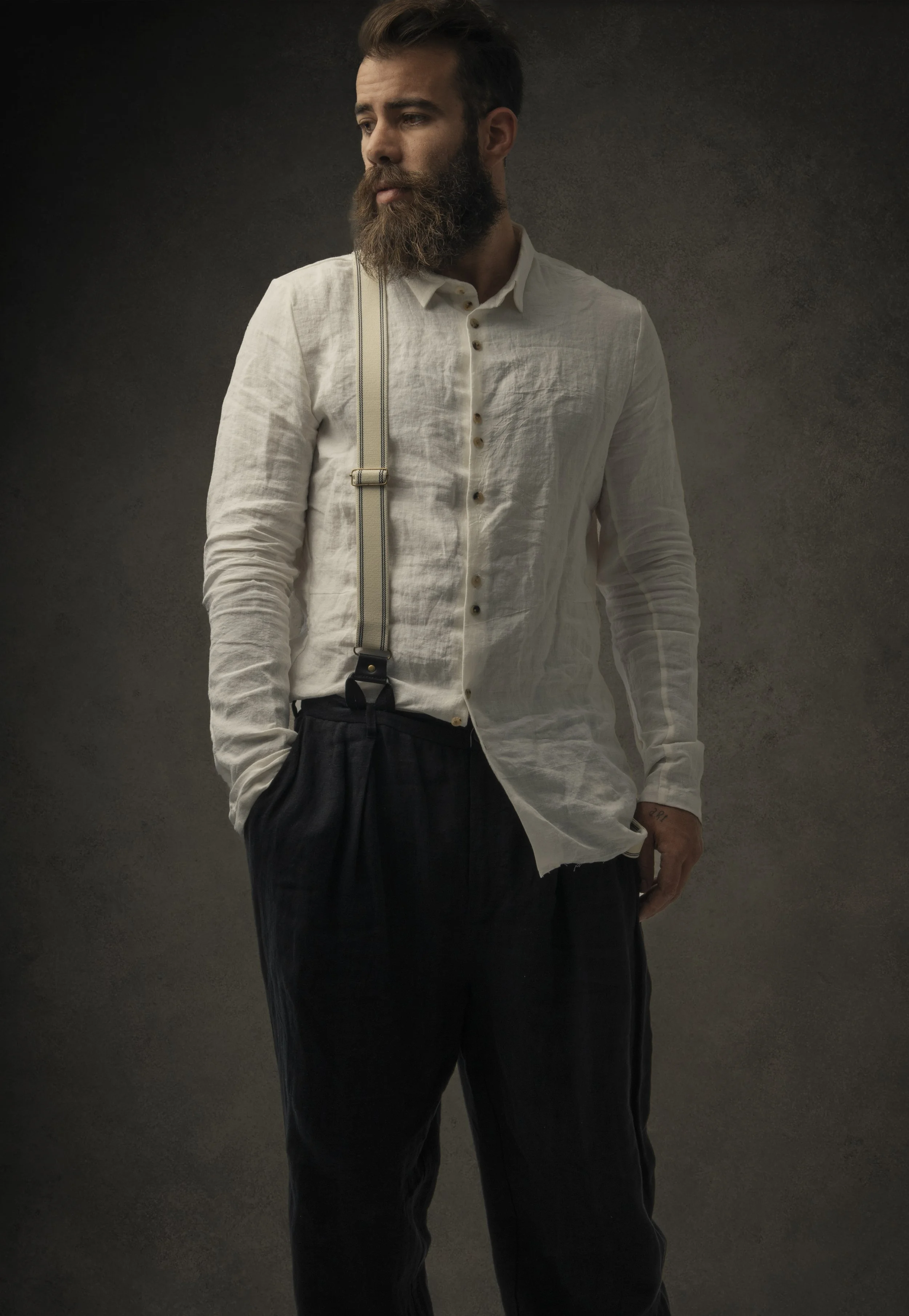A man with a beard and mustache looking to the side, wearing a white button-up shirt, black pants, and suspenders, standing against a dark background.